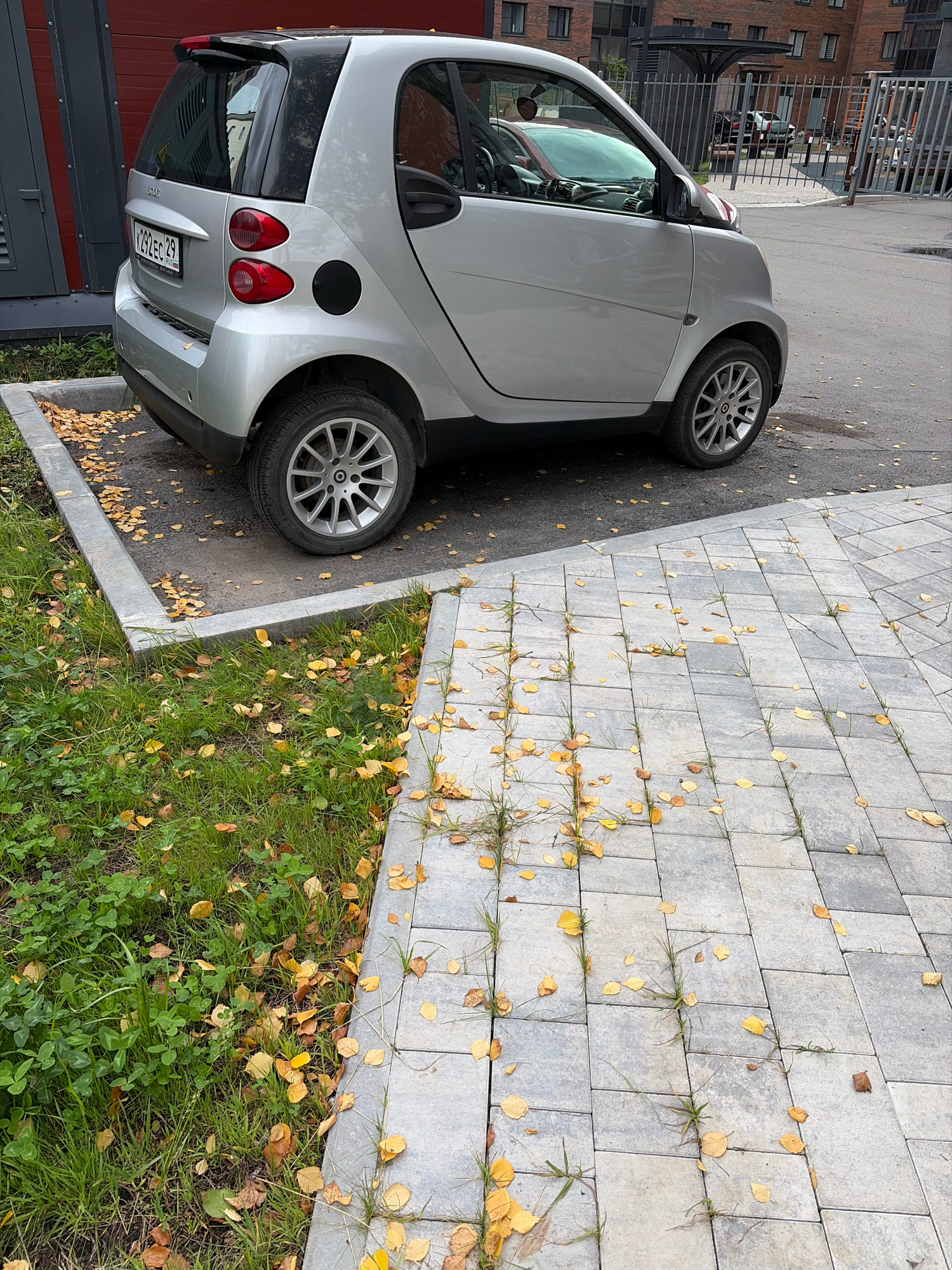 A silver Smart Fortwo car is parked in an outdoor paved area. The car is facing towards the right of the frame. The background shows a building with brickwork and a fence. The ground is a mix of asphalt and paving stones, with some grass and fallen yellow leaves visible. The scene appears to be in an urban setting, possibly a residential courtyard or parking area in Arkhangelsk, Russia, given the presence of a Russian license plate on the car. There are no people visible in the image.