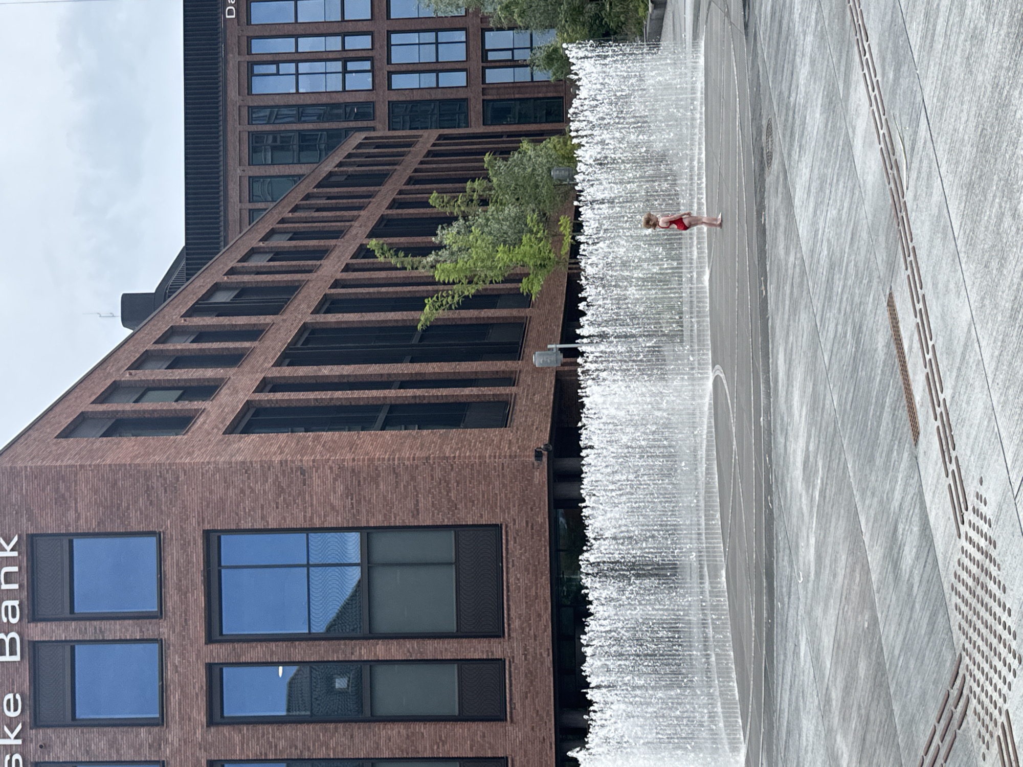 The image shows a young child wearing a red swimsuit standing in a water fountain in an urban plaza. The child is facing to the right, with their back slightly to the camera. The fountain consists of numerous jets of water spraying upwards and outwards in a semi-circular pattern, creating a shimmering curtain of water. Behind the fountain are modern brick buildings with large windows. One building has text that reads "Ske Bank" and another building has text that reads "D". The plaza pavement is made of large grey square tiles. The scene appears to be located in Copenhagen, Denmark, given the presence of "Ske Bank" signage. The sky is overcast.