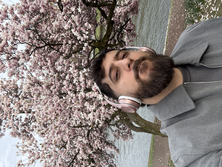 A male individual is depicted in a close-up selfie-style portrait, positioned outdoors on the bank of a river in Strasbourg, France. The man, with dark hair and a full dark beard, wears rose gold and white Beats Solo headphones. He is dressed in a grey collared jacket over a grey ribbed top. His expression is neutral as he looks directly into the camera. Behind him, a large magnolia tree is in full bloom, covered in numerous light pink and white blossoms. To the left and right of the tree's trunk, a calm body of water, likely the Ill River, is visible. Scattered light-colored petals are present on the ground along the riverbank in the foreground. The sky overhead is overcast and light grey.