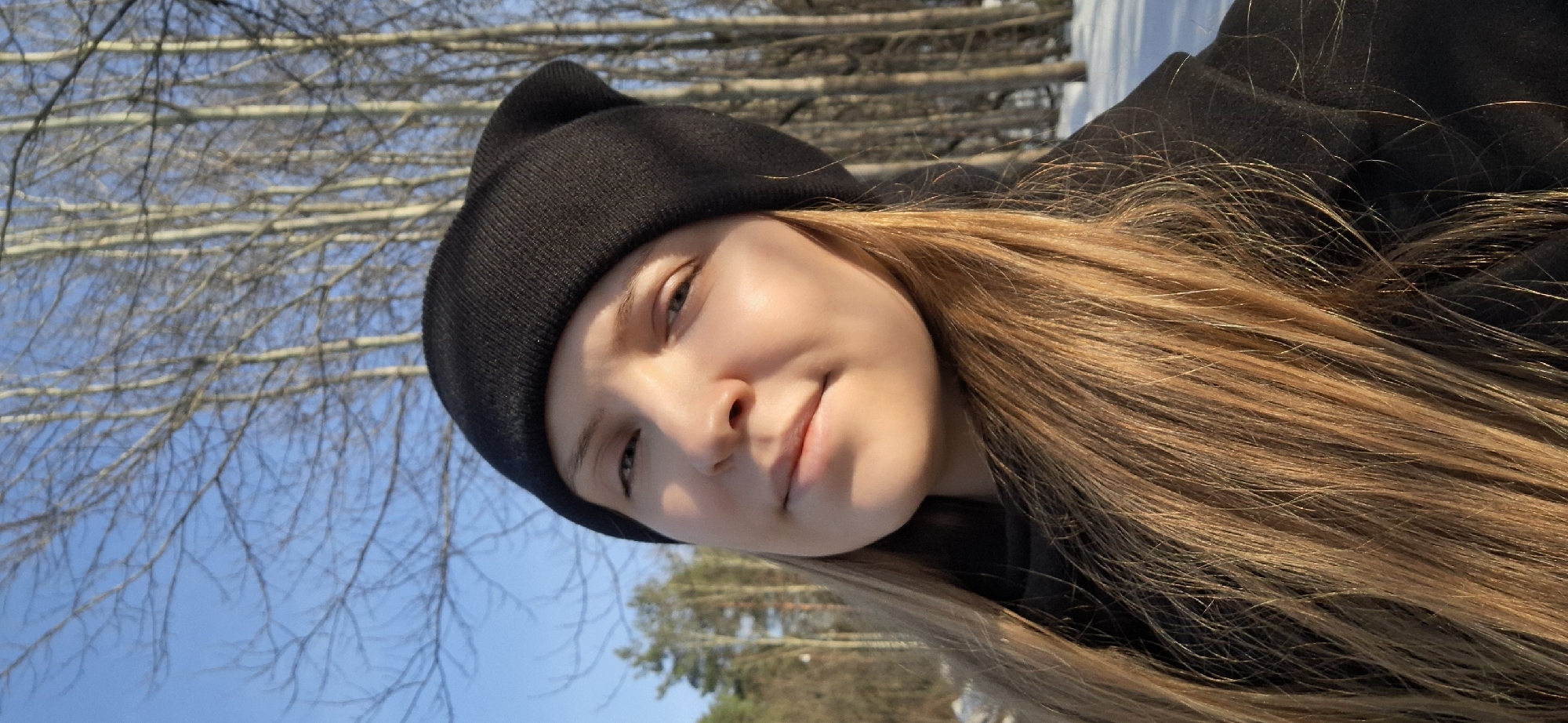 A young woman wearing a black beanie is taking a selfie outside in what appears to be a wooded area on a sunny day. The sky is clear and blue, visible between the bare tree branches in the background. The woman has long, light brown hair. There are other trees and some snow on the ground. The location may be Chaikovsky, Russia.