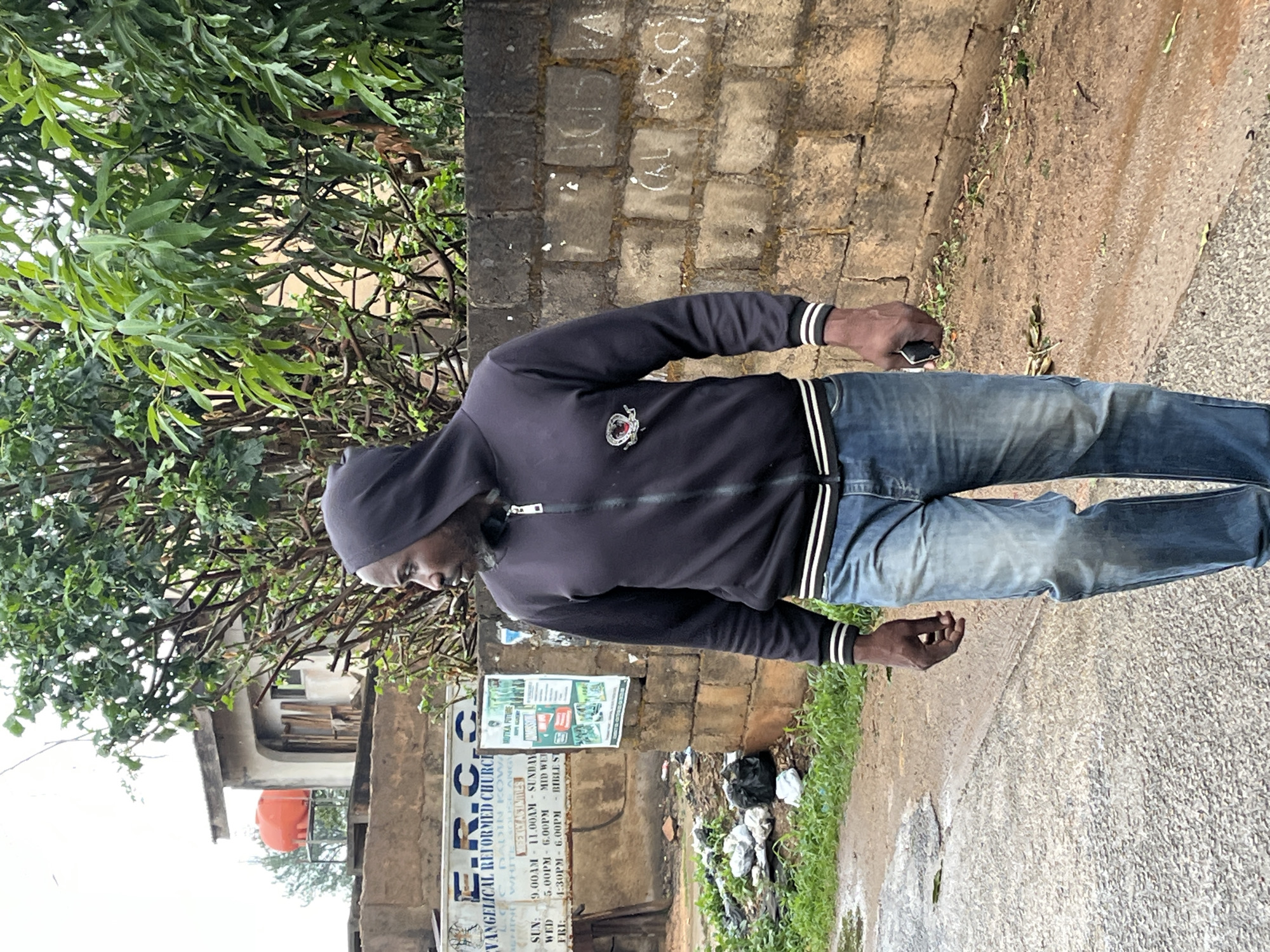 The image depicts a man standing on a paved road in Jos, Nigeria. The man is wearing a dark hooded jacket with white stripes on the sleeves, jeans, and a dark hood over his head. He is holding a small dark object in his right hand. Behind him is a weathered brick wall, partially obscured by greenery. A sign for the Evangelical Reformed Church (E.R.C.C) Adin Kow is visible. There's trash along the base of the wall. A building is seen in the background to the left of the sign.