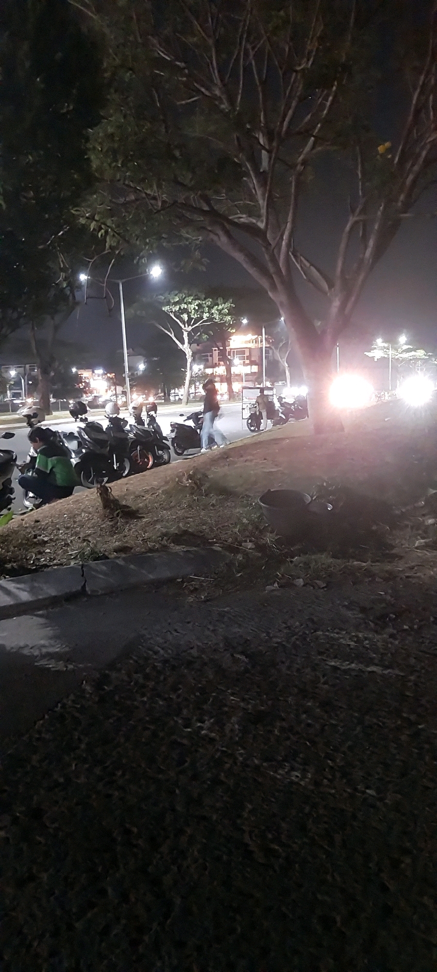 The image depicts a nighttime scene on a street in Medan, Indonesia. In the foreground, a grassy embankment is visible, with a dark-colored bucket to the right. Beyond the embankment, a row of parked motorcycles is lined up along the side of a street. One person, wearing a dark jacket and light-colored pants, is walking away from the motorcycles towards the right of the frame. Another individual, wearing a green top, is seated on one of the motorcycles. In the background, streetlights illuminate the scene, and buildings with visible lights are present. Trees with bare branches are also scattered throughout the background. The overall atmosphere suggests an urban environment at night.