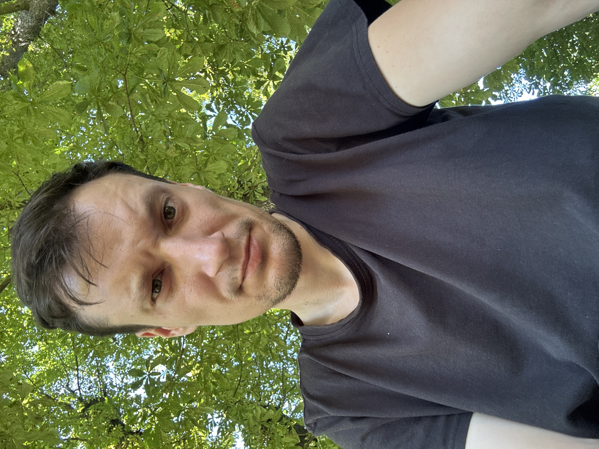 A man with short, dark hair, a light beard, and fair skin is pictured from the chest up. He is wearing a black t-shirt. His face is turned slightly towards the camera, and he appears to be looking directly at the viewer with a neutral expression. Behind him, a bright green canopy of leaves from a large tree is visible against a bright sky. The image was taken outdoors in Makiv, Ukraine, during daylight hours, likely on a sunny day given the lighting.