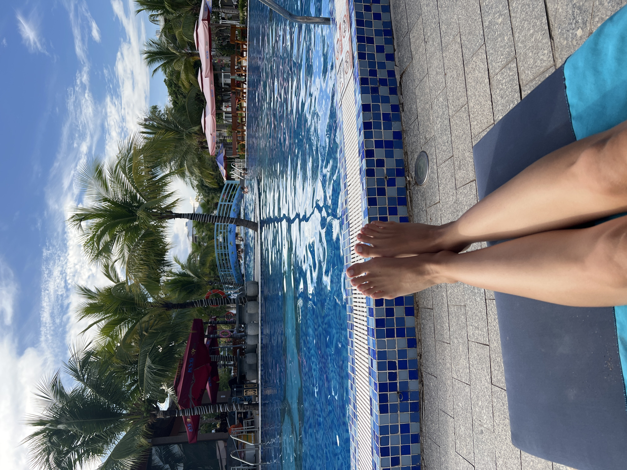 The image depicts a person's legs stretched out on a blue lounge chair next to a swimming pool in Nam Nha Trang Ward, Vietnam. The pool is lined with blue and white tiles. Palm trees and umbrellas are visible in the background against a partly cloudy sky.