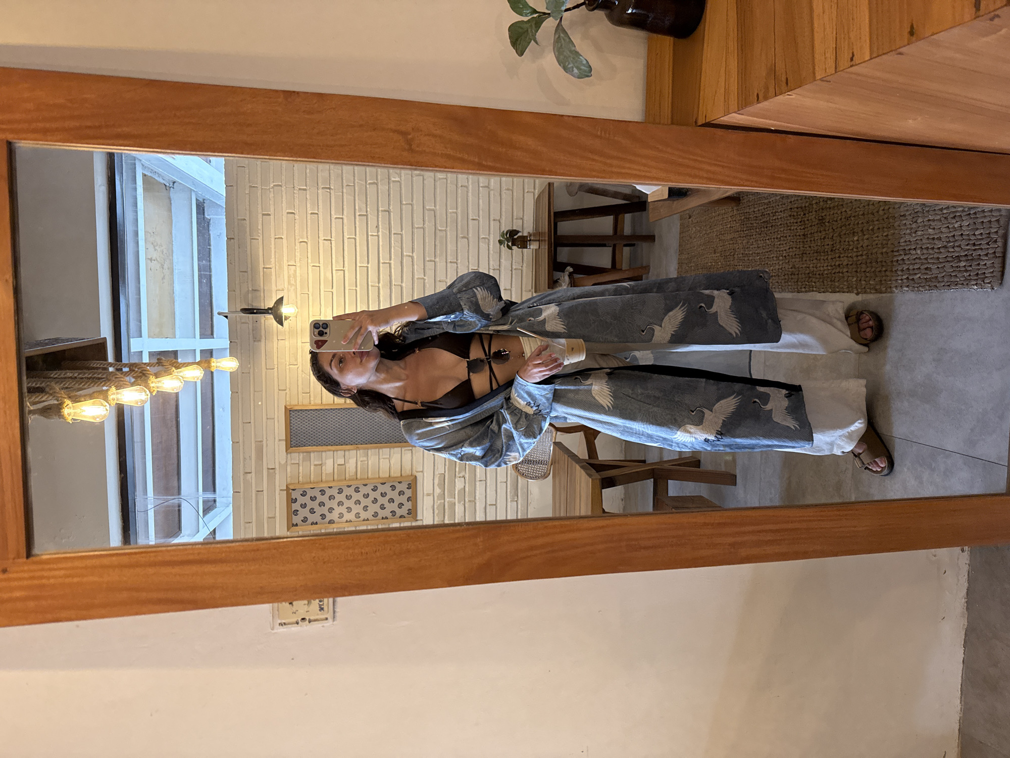 A woman is standing in front of a mirror in an indoor setting in Unknown, Indonesia, taking a photo of herself. The woman has dark hair and is wearing a black bikini top, white pants, and a blue kimono-style robe with a crane pattern. She is holding a smartphone with her right hand, taking a mirror selfie.
The background includes a white brick wall with two decorative panels, a wooden chair, and a rug. The mirror's wooden frame is visible on the edges of the image. The lighting is a combination of natural light and artificial light from a hanging fixture. The scene appears to be a stylishly decorated interior.