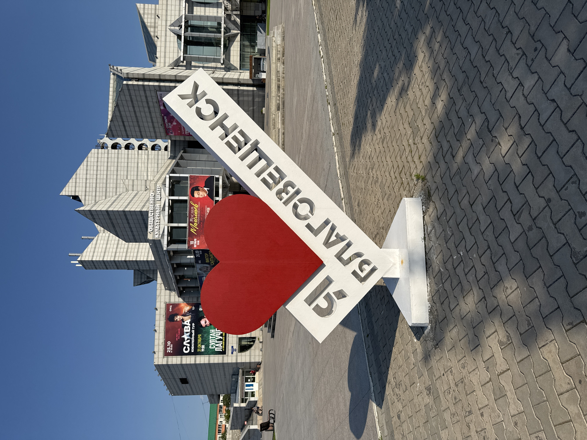 A wide shot captures an outdoor plaza in front of a modern, multi-faceted white building complex, under a clear blue sky. In the foreground, a large, stylized sculpture forms the centerpiece: a bright red heart on the left, and a white, angled structure with cutout letters on the right. The white structure reads "Я БЛАГОВЕЩЕНСК," which translates to "I Love Blagoveshchensk." The sculpture is positioned on a patterned gray pavement.
Behind the sculpture, the building complex features a modern architectural style with numerous glass panes, angled white panels, and protruding sections. Several large advertisements are mounted on the building's facade, displaying images of performers and event dates. To the left of the main building, a smaller, light-colored building with a green roof is visible.
In the middle ground, a wide, paved plaza stretches out, with shadows from the sculpture and unseen objects cast across it. A few benches and a set of stairs are visible on the left side of the plaza. A few people are seen seated on the benches in the distance. The overall environment suggests a public space in a city, likely a cultural center or a prominent landmark. The context provided by the sculpture explicitly identifies the location as Blagoveshchensk, Russia.