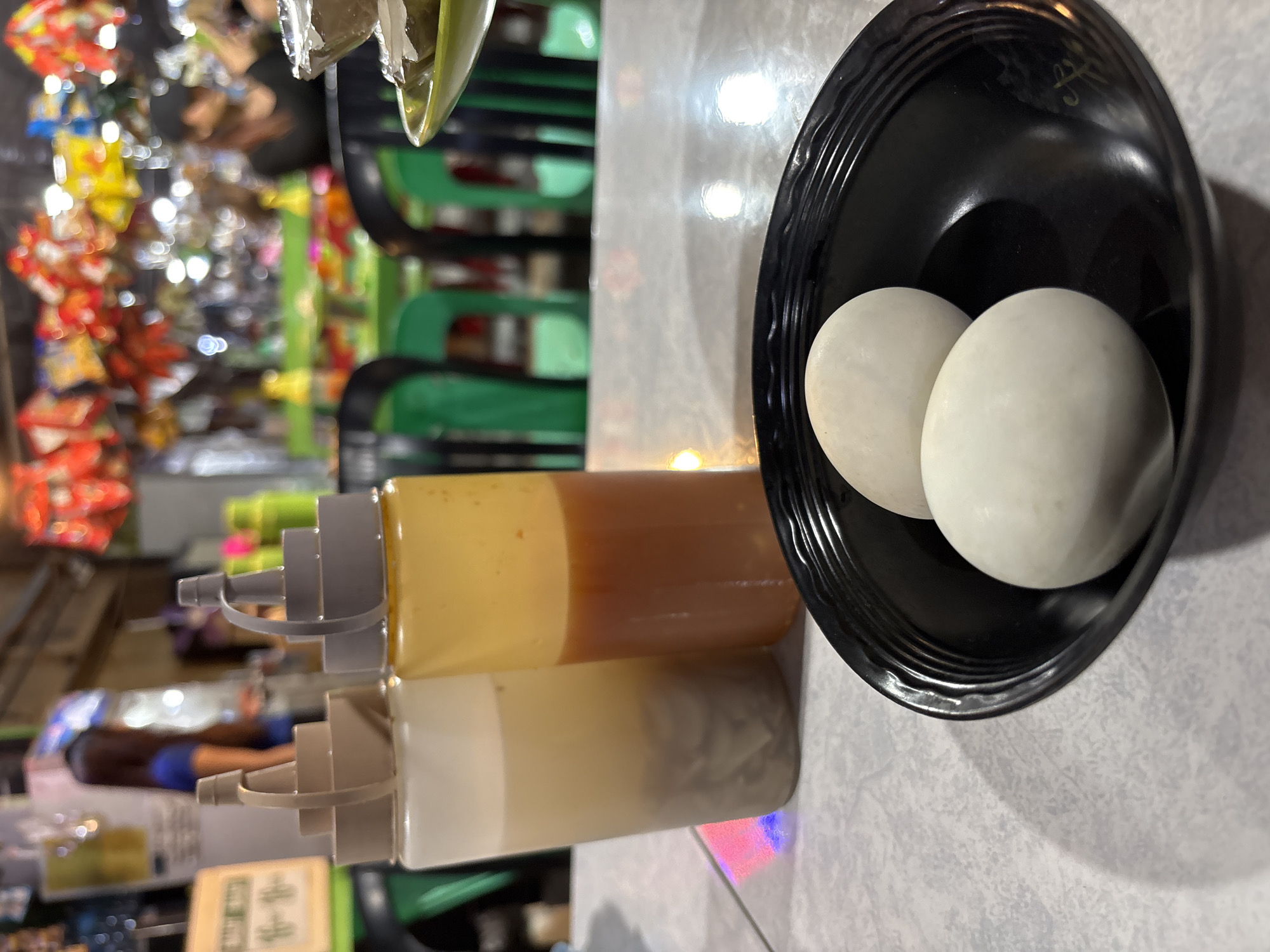 This image captures a close-up view of a table setting at what appears to be an outdoor eatery in Carmen, Philippines. In the foreground, a black, ridged bowl holds two pale, oval objects, presumably duck eggs, which are a common street food in the Philippines. To the left of the bowl are two condiment bottles: one filled with a light-colored liquid containing what seems to be pickled vegetables or slivers of meat, and another filled with a reddish-brown sauce, possibly a chili sauce or vinegar-based condiment. The bottles have gray and beige stoppers. The table surface has a speckled pattern. In the blurred background, several green plastic chairs surround tables, and the scene suggests a busy public space with colorful decorations hanging overhead and various individuals in the distance, one of whom is wearing a black cap. The lighting is a mix of artificial and natural, creating a lively atmosphere typical of a local market or food stall.