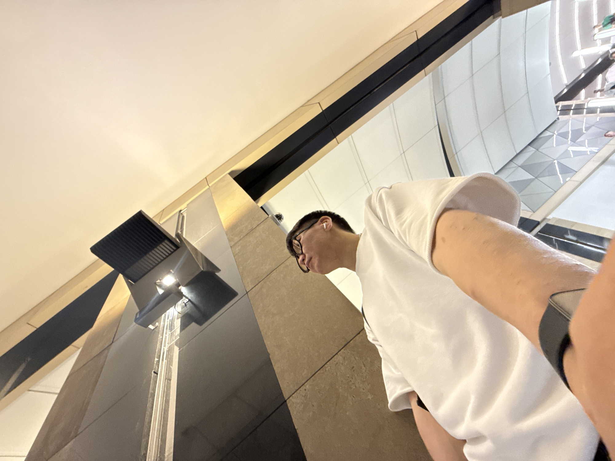 An upward-angle, waist-up shot shows a young East Asian person wearing glasses, a white t-shirt, and white wireless earbuds. The person is looking up and to the left. Their left arm is extended towards the camera, and a black smartwatch is visible on their wrist. The background consists of a modern, indoor architectural space with polished stone walls and a white ceiling with a grid pattern. A light fixture is mounted on the wall, and parts of an escalator and some distant figures are visible in the background. The lighting is soft and ambient, with a warm glow from the ceiling. The scene appears to be a public indoor space, possibly a shopping mall or transportation hub. There is no indication of the specific location being in Moscow, Russia.