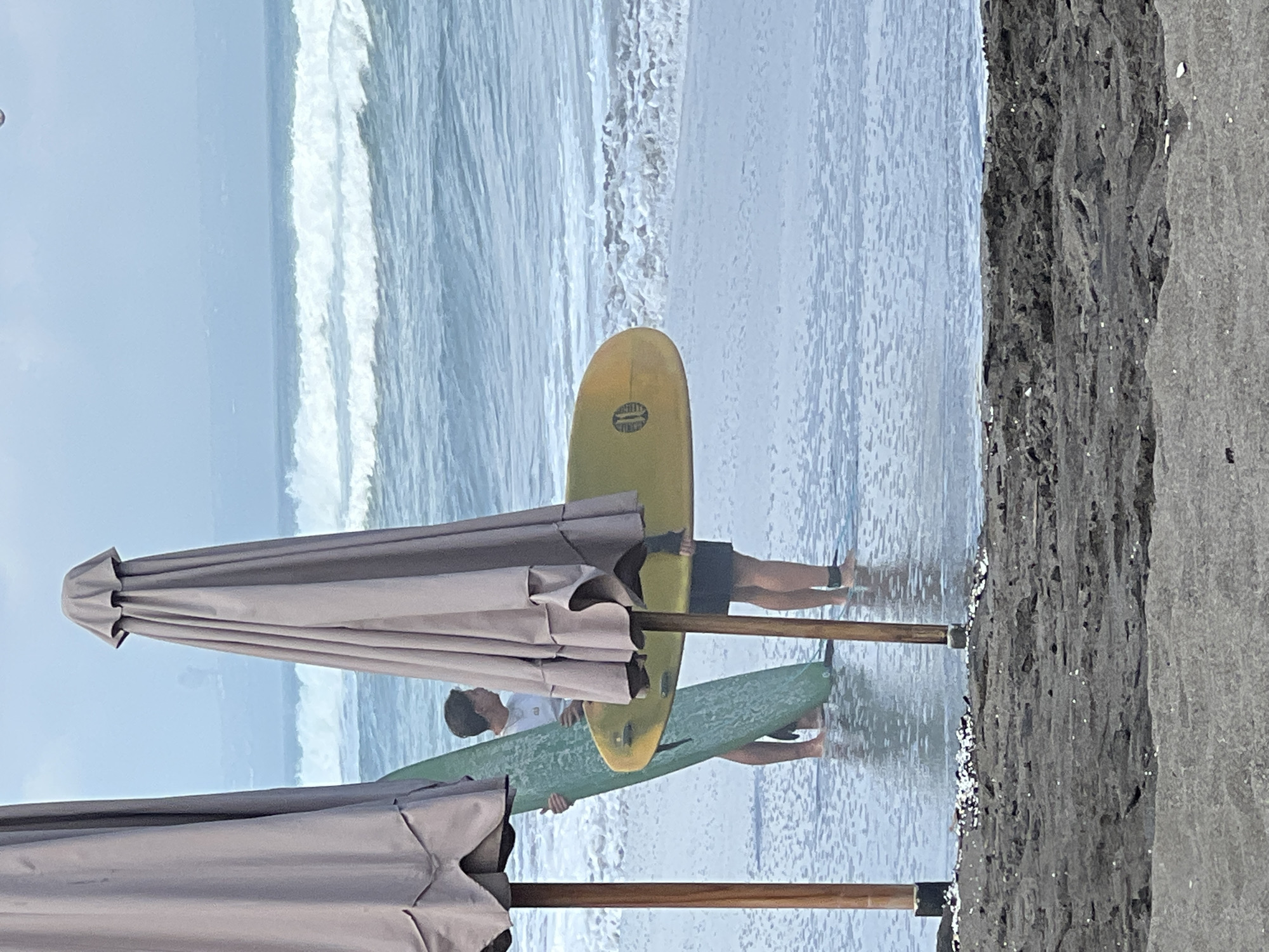 The image shows a beach scene in North Kuta, Indonesia, with two people standing in shallow water near surfboards and beach umbrellas. The ocean has gentle waves, and the sky is overcast. The ground is dark sand with some debris. One person is holding a green surfboard, and the other is holding a yellow surfboard. Both individuals are wearing swim trunks. The beach umbrellas are large and light-colored, providing shade.