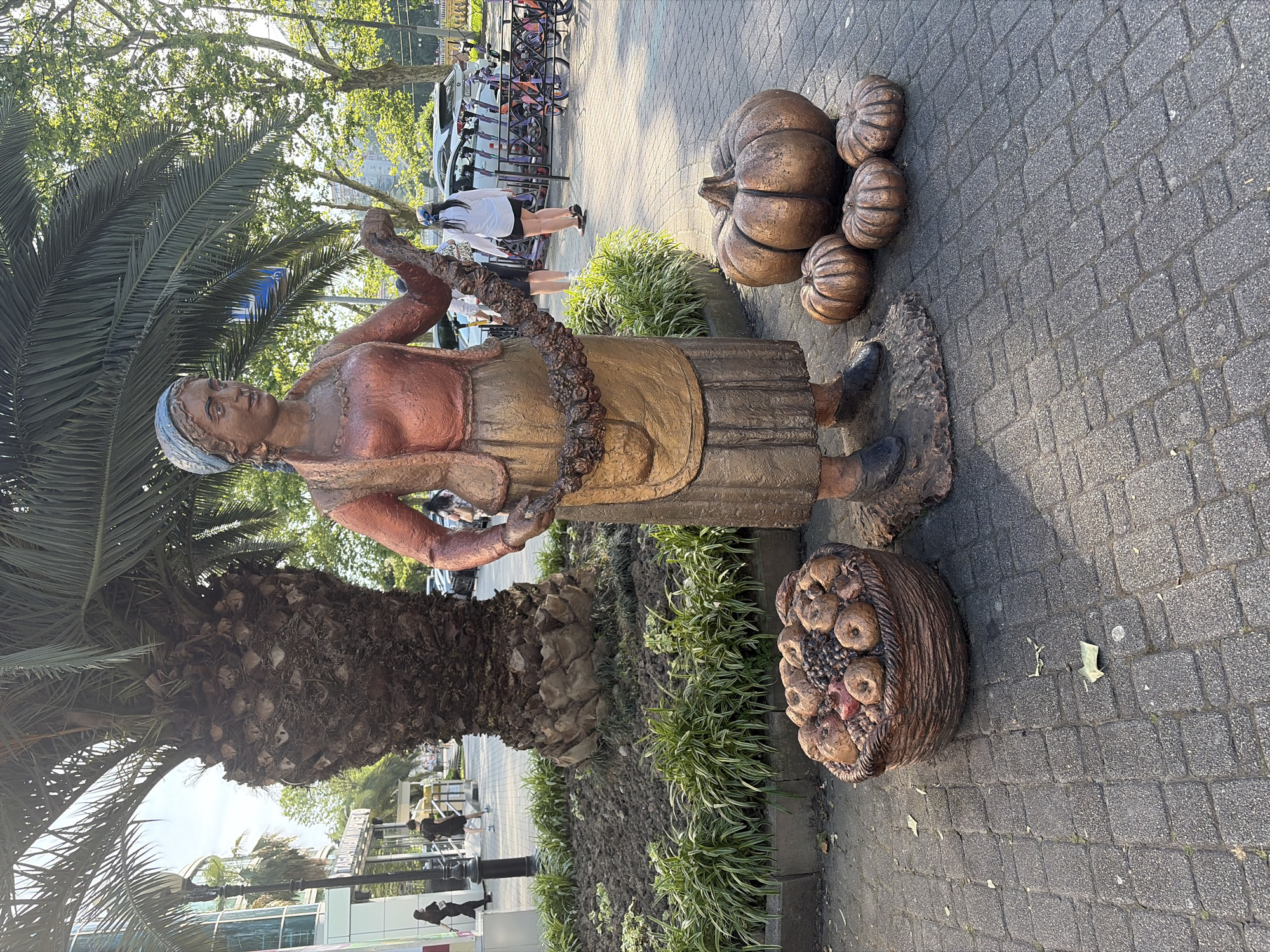 The image depicts a bronze-colored statue of a woman with accompanying bronze-colored fruit and vegetables, situated on a pedestrian paved area in Sochi, Russia.
The central subject is a standing statue of a woman, possibly representing a market vendor or farmer. She wears a blue headscarf, a reddish-orange top, a long gold-colored skirt or apron, and dark shoes. Her left arm is bent, holding a garland of what appears to be dried fruit or flowers, draped across her torso.
To the left of the statue's base, a bronze-colored basket filled with various fruits, including apples, grapes, and pomegranates, rests on the pavement. To the right of the statue, one large and three smaller bronze-colored pumpkins or gourds are arranged on the ground.
The statue and these items are positioned on a grey paver block walkway. Behind the statue, a raised plant bed contains dark soil and low-lying green and white variegated plants.
In the mid-ground, a large palm tree with a thick, textured dark brown trunk and prominent green fronds dominates the center. To the left of the palm tree, a modern building with large windows and light-colored siding is visible. Part of a sign in Cyrillic, "Табачный мир" (Tobacco World), is discernible on the building. A black lamppost stands near the building.
Several pedestrians are present in the background. On the left, two individuals in dark clothing walk away from the camera. Another person in dark clothing walks past the palm tree. On the right, a woman in a white shirt and black shorts walks away from the camera, near parked vehicles and a rack holding multiple purple shared electric scooters and bicycles.
The background on the right features numerous green deciduous trees. The lighting suggests strong sunlight, casting shadows towards the lower left, indicating a clear day, likely mid-morning or late afternoon.