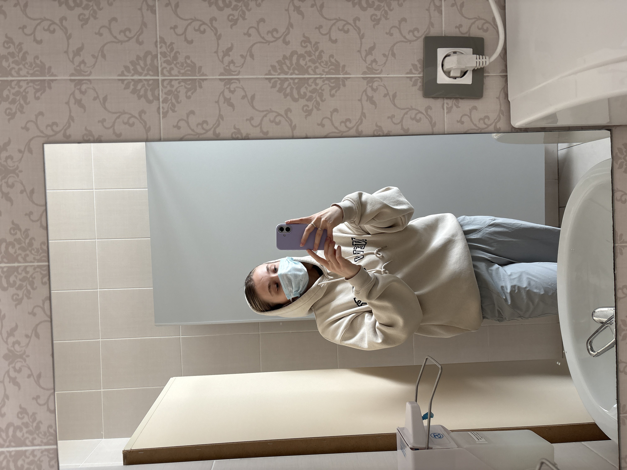 A young person wearing a face mask is taking a selfie in a mirror in what appears to be a bathroom. The person is wearing a hooded sweatshirt and light-colored pants. The background includes tiled walls with a floral pattern, a white sink, a soap dispenser, and an electrical outlet. The location is Izhevsk, Russia.