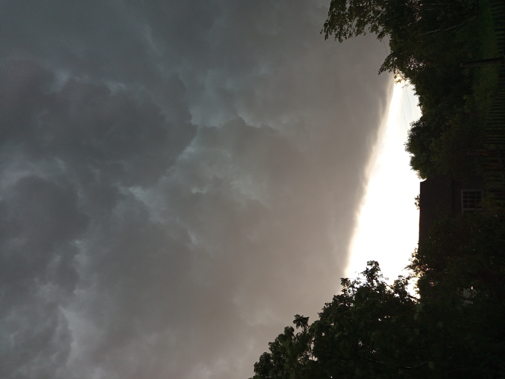 The image depicts a dark, cloudy sky over a rural landscape, likely in Ostrovtsy, Russia, as suggested. The sky is dominated by heavy, grey storm clouds, with a band of brighter light peeking through near the horizon. In the foreground, silhouettes of trees frame the scene on the left and right. Below the sky, the tops of trees and a portion of a building with a red roof and a window are visible. A wooden fence runs along the bottom of the image. The overall atmosphere is somber and suggests an approaching storm.