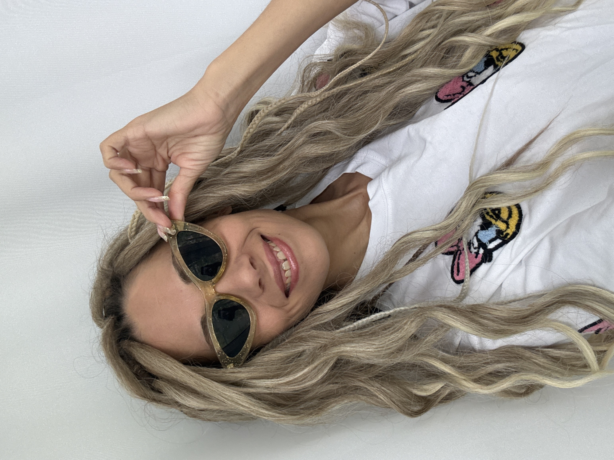 A light-skinned woman with blond dreadlocks is lying down, wearing a white t-shirt with patches on it, and holding up gold-framed sunglasses. The background appears to be a plain white surface. The location in the city of Rusanovo, Russia, cannot be determined from the image.