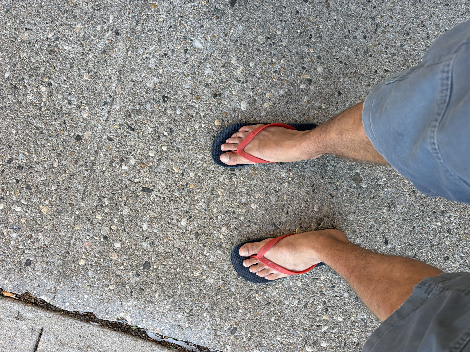 An overhead, eye-level shot shows a pair of bare feet in blue and red flip-flops, standing on a concrete sidewalk. The sidewalk has small pebbles embedded in it, and to the left, there is a grassy verge with a cigarette butt on it. The person is wearing grey shorts. The image was taken in Riverhead, United States of America.