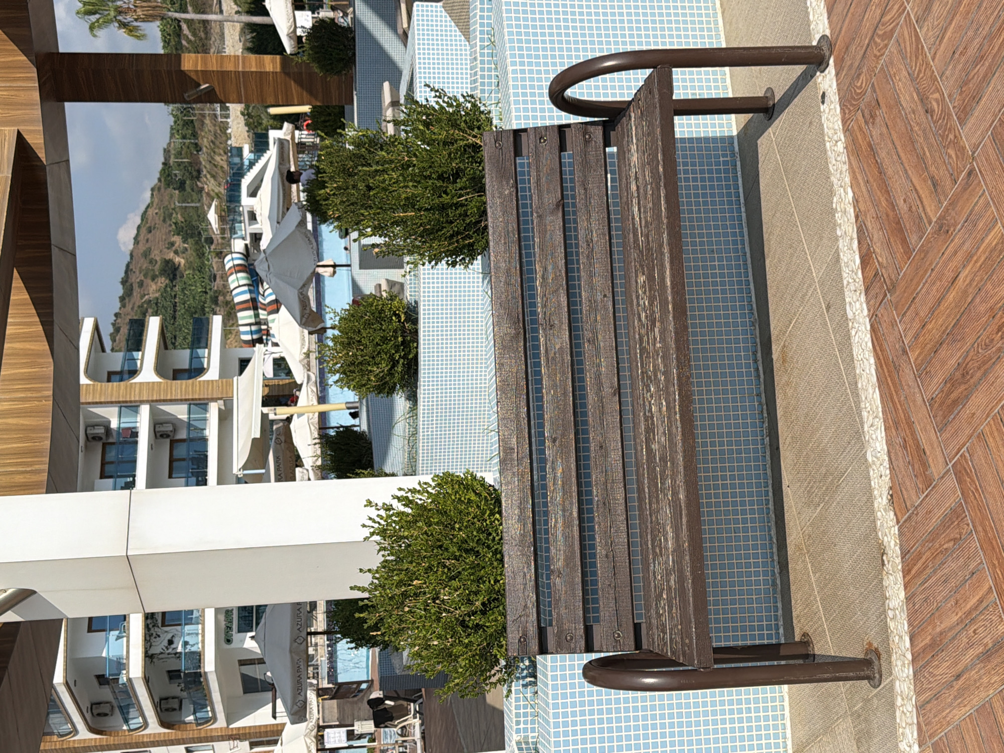 The image depicts a park or resort setting in Alanya, Turkey, featuring a wooden bench in the foreground. Behind the bench, a swimming pool area is visible with light blue tiled edges, surrounded by lush green bushes and white umbrellas. In the background, a modern white building with multiple balconies and air conditioning units is prominent, with a hillside dotted with trees and a palm tree to the right. A water slide is also visible in the distance. The ground is paved with wooden-patterned tiles.