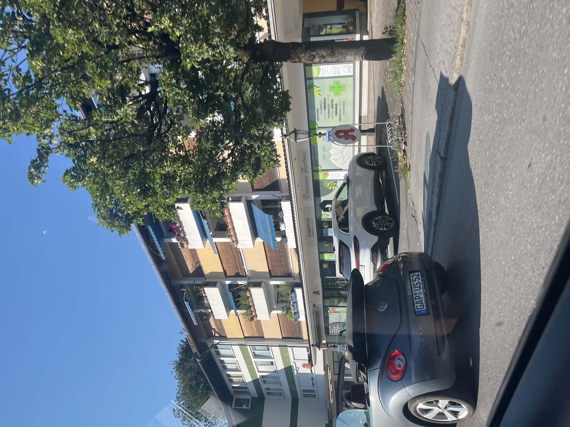 The image shows a street scene in Garmisch-Partenkirchen, Germany, on a sunny day. In the foreground, a grey Volkswagen Beetle is parked. Behind it, a white car is also parked. The street is lined with buildings, one of which is a pharmacy named "Dreitorspitz Apotheke." The pharmacy has a green cross logo and text in multiple languages, including German, English, and Polish, indicating it serves a diverse clientele. Above the pharmacy, residential floors with balconies decorated with plants and flowers are visible. A large tree with lush green foliage casts a shadow on the sidewalk and street. The sky is a clear blue, suggesting good weather. The overall impression is a typical European street scene with businesses and residences coexisting. The license plate of the Volkswagen Beetle reads "GAP US 52," with "GAP" likely referring to the district of Garmisch-Partenkirchen.