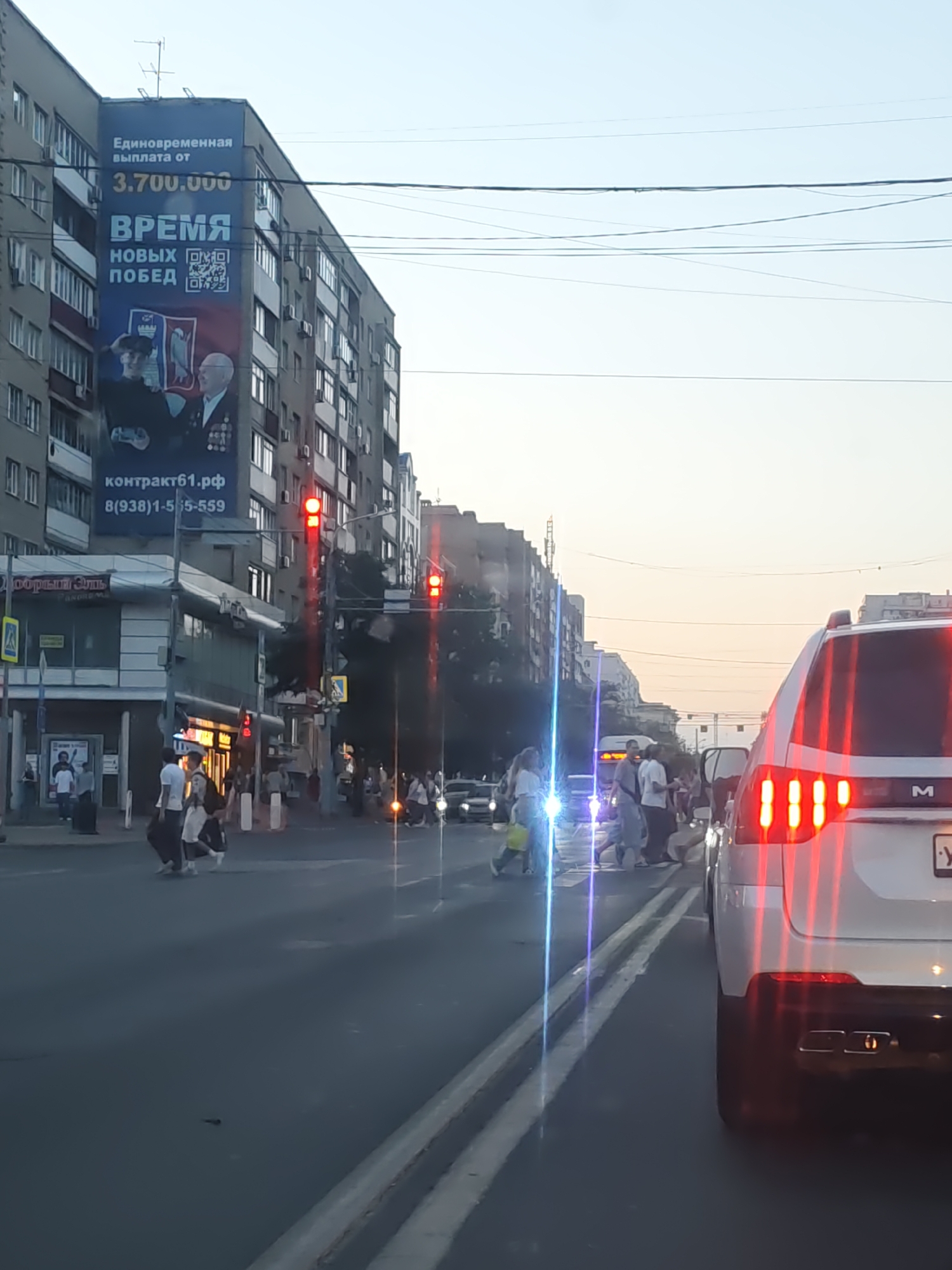 The image depicts a street scene in Rostov-on-Don, Russia, during what appears to be early evening or dusk. A large billboard on the side of a multi-story apartment building is prominent, advertising "Единовременная выплата от 3.700.000" (One-time payment from 3,700,000), followed by "ВРЕМЯ НОВЫХ ПОБЕД" (Time for new victories). The billboard also includes images of two men and a QR code.
Below the billboard, a commercial building with glass windows and signage is visible. The street is busy with pedestrian and vehicular traffic. Several traffic lights are illuminated red, indicating a stop. People are crossing a pedestrian intersection. On the right side of the image, the rear of a white SUV is visible, its brake lights illuminated. The overall environment suggests an urban setting with buildings, streetlights, and infrastructure.