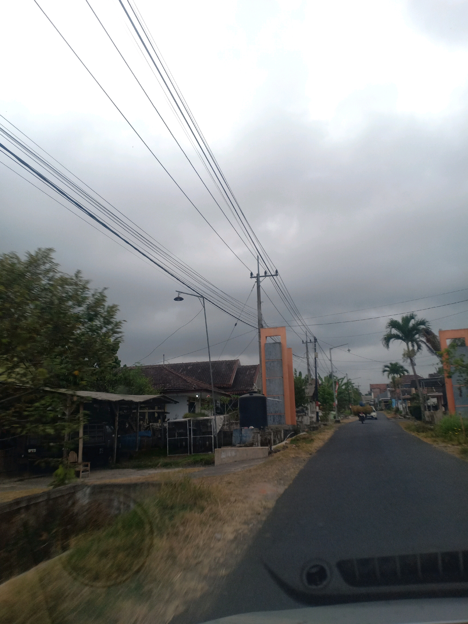 The image depicts a cloudy day scene on a paved road in Kloposawit, Indonesia. The road runs through a residential area with houses and buildings on either side. Power lines crisscross the sky, supported by utility poles. To the left of the road, there are trees and a covered structure with a vehicle parked underneath. Further down the road, a motorcycle carrying a load of hay is visible, as well as a small white car. The overall impression is of a quiet, rural or semi-urban environment under an overcast sky.