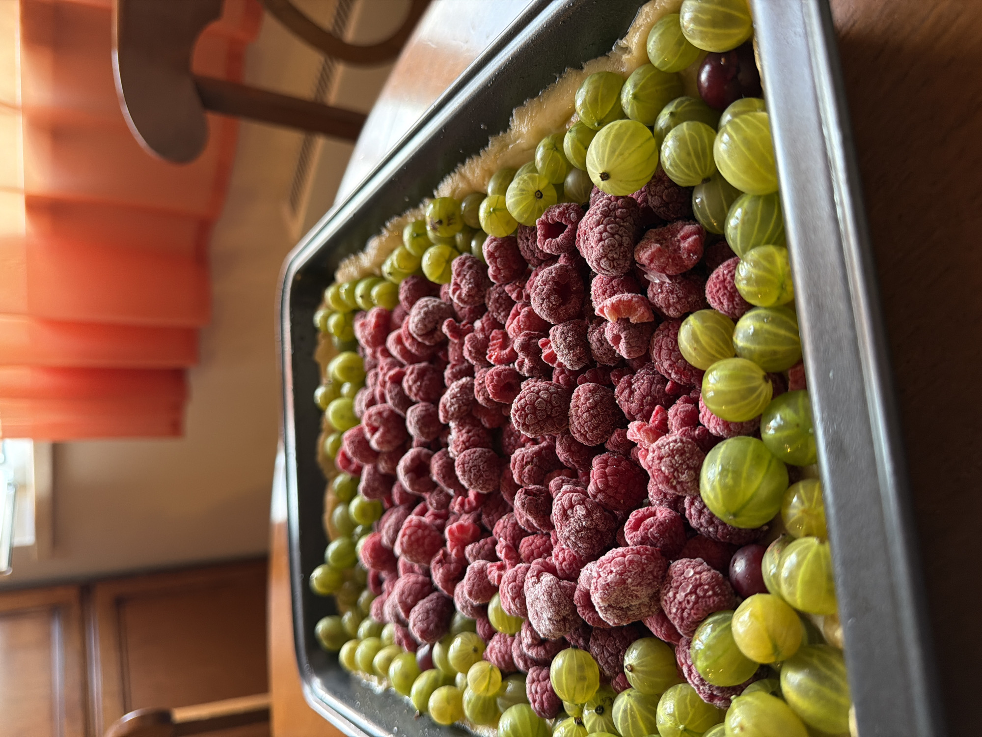 The image shows a close-up, eye-level view of a rectangular baking pan filled with a mixture of frozen raspberries and gooseberries, arranged on a crust. The berries are clustered in the pan, with raspberries forming a central mass and gooseberries arranged around the edges. The background is blurred, but suggests an indoor setting with wooden furniture and orange curtains. There is no indication of Moscow, Russia, or any specific city in the image.