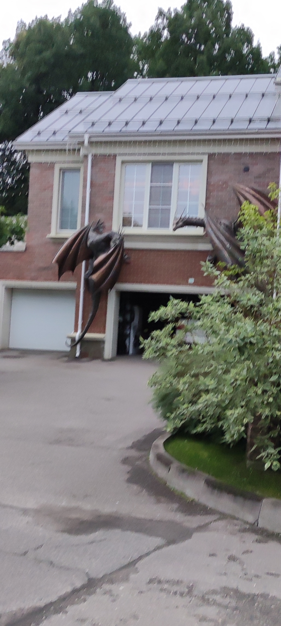 The image depicts a brick building with a metal roof, likely a residential or commercial property, in Rosinka, Russia. Two large, metallic dragon sculptures are prominently displayed on the exterior of the building. One dragon is positioned on the left side, appearing to cling to the brickwork with outstretched wings. Another dragon is visible on the right side, its head emerging from a window or opening. In front of the building, a paved driveway leads to a garage door. The environment appears to be outdoors, with trees visible in the background and foliage near the right side of the building. The ground in the foreground shows signs of wetness, possibly from rain.