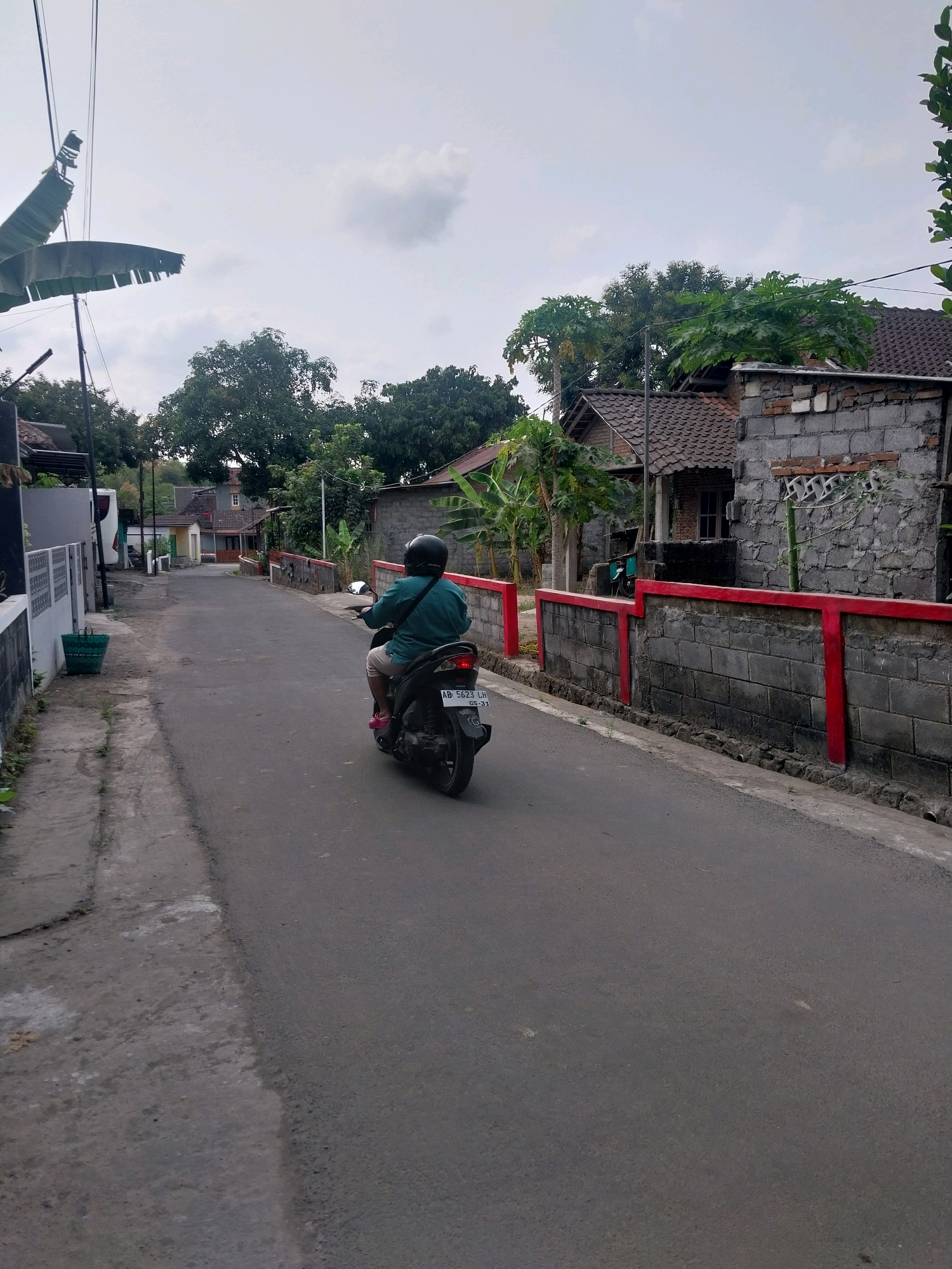 A person wearing a teal shirt and a helmet is riding a black scooter down a paved road in Purwomartani, Indonesia. The scooter has a license plate that reads "AB 5623 LH". The road is lined with houses on both sides, some with tiled roofs and others with concrete walls. A red-painted concrete wall with vertical red supports runs along the right side of the road. Trees and vegetation are visible throughout the scene, with a few power lines overhead. The sky is overcast with some clouds.