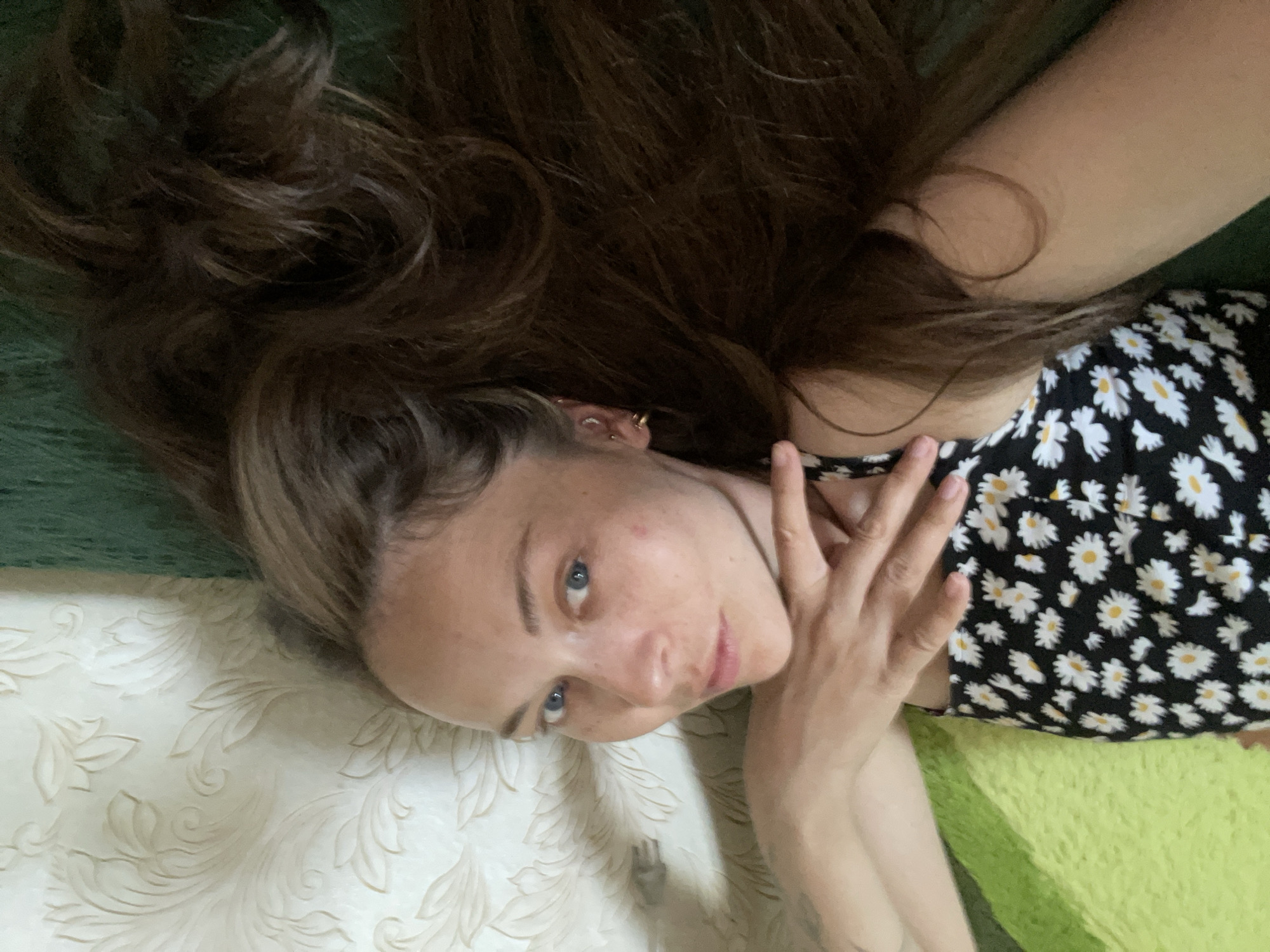 A young woman with long, brown hair and blue eyes is lying on her back on a green surface. She is wearing a black dress with a white daisy pattern. Her left arm is bent and her hand is resting on her face. The background is a patterned wallpaper. The scene appears to be indoors, possibly in a bedroom or living room.