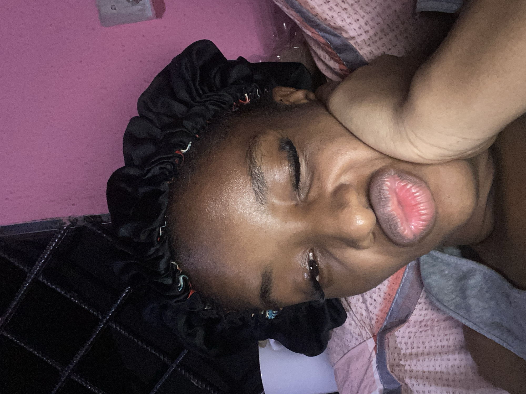 A dark-skinned young woman wearing a black satin bonnet is lying in bed, making a kissing face. Her left eye is open, and her right eye is closed, with visible eyelashes. Her hand is resting on her cheek. The background includes a pink wall and what appears to be a metal grate structure. The bedding is pink and patterned. The scene is indoors, likely in a bedroom, and could be located in Jos, Nigeria, given the prompt's context.