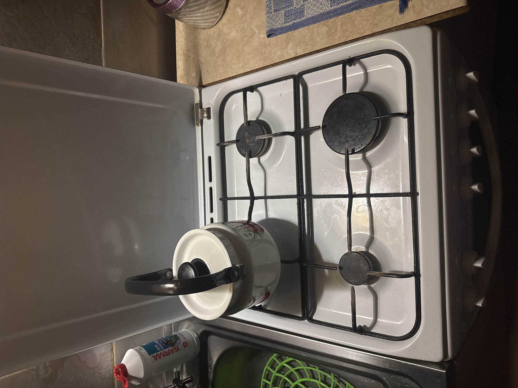 A white gas stove is shown in a kitchen, with a white enamel kettle with a red floral design placed on the left burner grate. To the left of the stove is a stainless steel sink with a green plastic drainer. A bottle of SARMA dishwashing detergent is visible next to the sink. The stove has four black burners and a white cooktop. The stove's lid is open, revealing the burners. The background shows a tiled wall and a countertop.