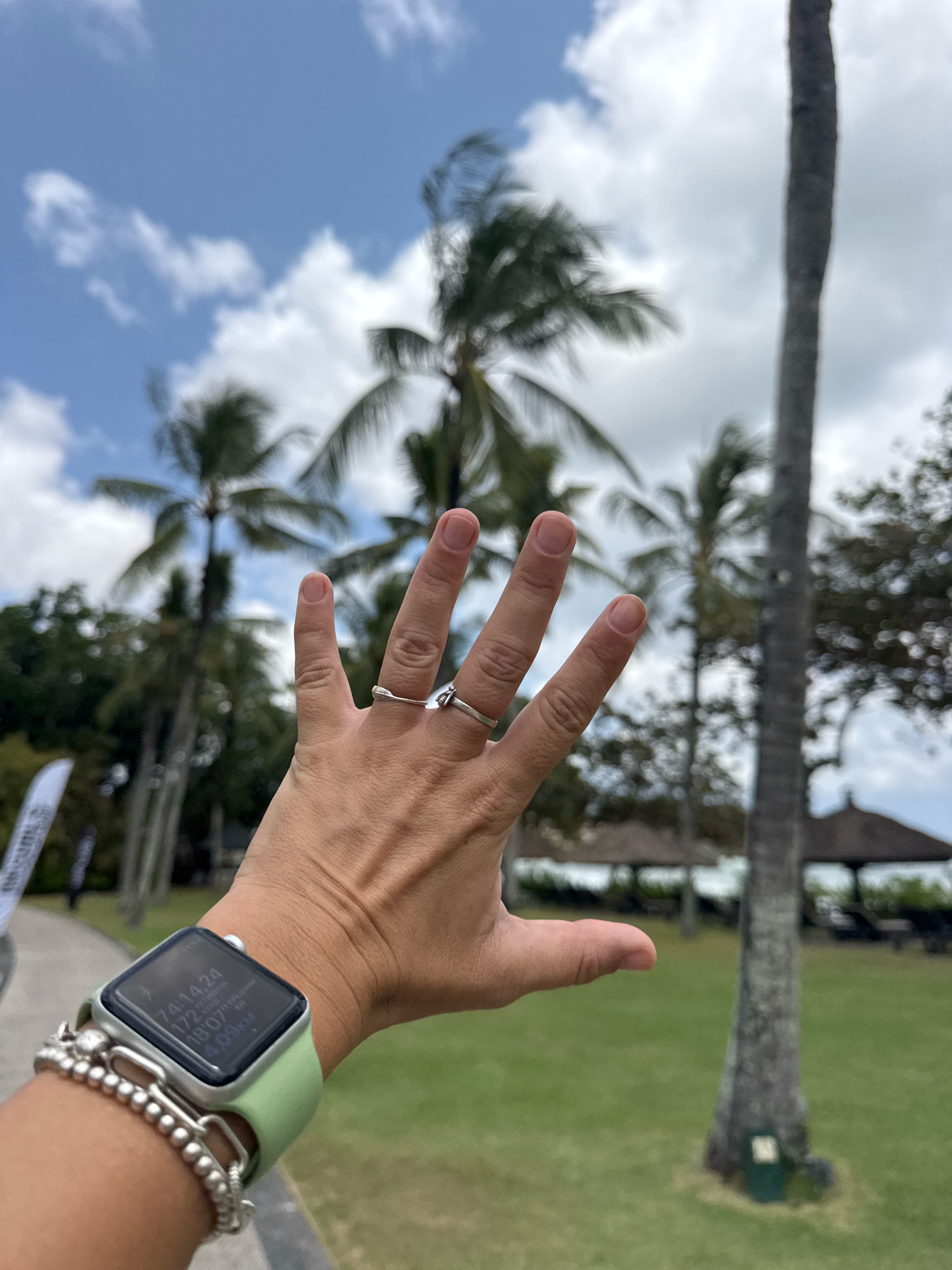 A person's left hand is held up with fingers splayed in the foreground of the image. The person is wearing a silver ring on their ring finger and another ring on their middle finger. A light green smartwatch is on the person's left wrist, displaying fitness metrics. The background shows palm trees, lush green grass, and a blue sky with scattered clouds. In the distance, there are thatched-roof structures and lounge chairs, suggesting a resort or outdoor recreational area. The setting appears to be Kuta Selatan, Indonesia, given the tropical vegetation and architectural style.