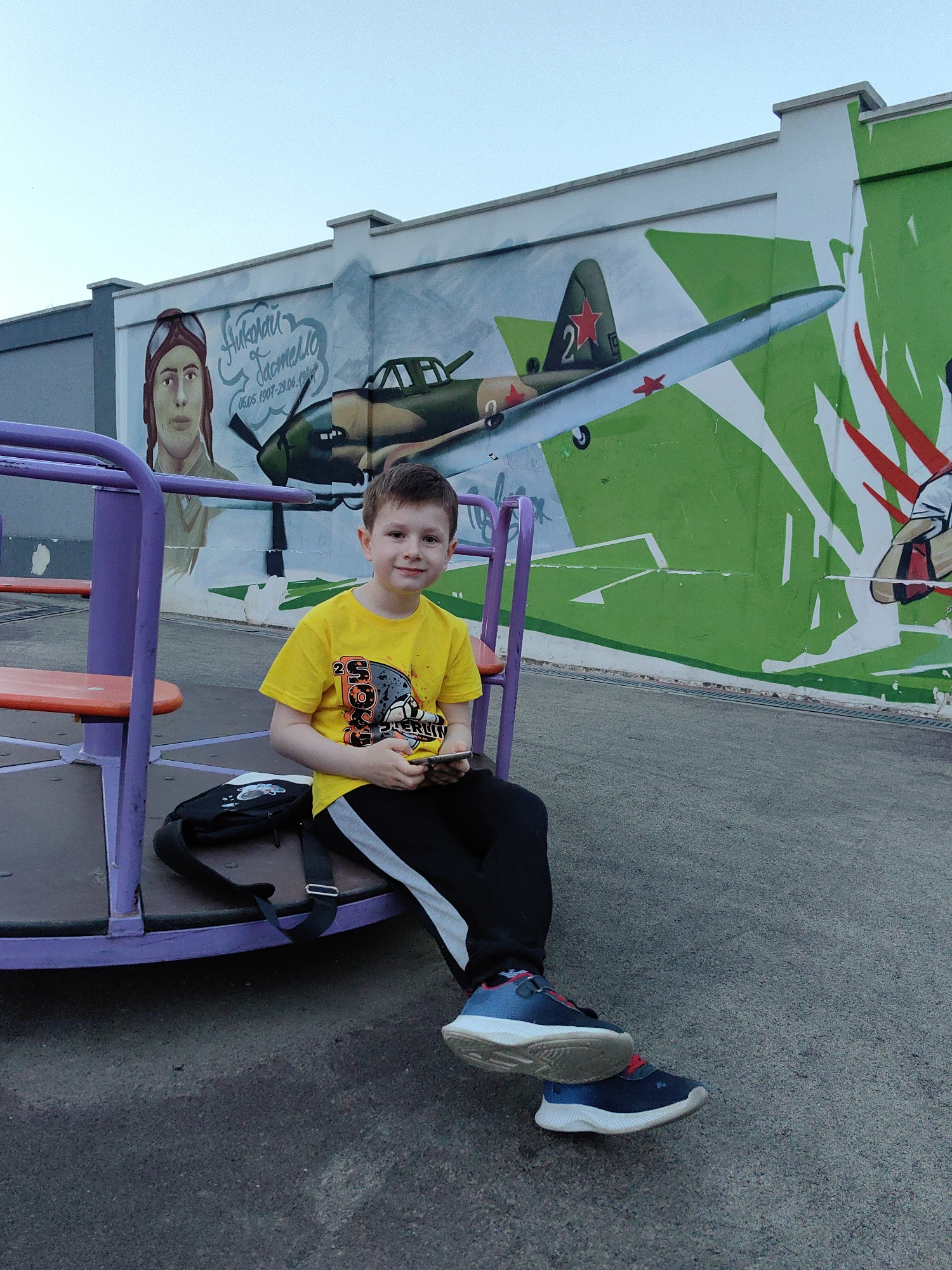 This image captures a young boy seated on a merry-go-round in an outdoor setting in Sochi, Russia. The boy, approximately 6-8 years old, has short brown hair and is wearing a yellow t-shirt with a "20 SPORTS WORLD" graphic, black track pants with grey side stripes, and blue and red athletic shoes. He is holding a smartphone in both hands and smiling directly at the camera. A black shoulder bag with a white graphic design rests beside him on the merry-go-round's dark platform.
Behind the boy, a large mural painted on a building wall depicts a historical theme. On the left side of the mural, a portrait of a male pilot wearing a flight helmet and goggles is visible. Adjacent to the portrait, Cyrillic text reads "Николай Гастелло" (Nikolai Gastello) followed by dates "06.05.1907-26.06.1941." To the right, a military aircraft, likely an Ilyushin Il-2 or similar, with camouflage paint (green and brown) and a red star with the number "2" on its tail fin, is shown in flight. The mural's background transitions from a light blue sky on the left to abstract green and white geometric shapes on the right. The ground is paved with dark asphalt. The overall scene suggests a public park or playground area with commemorative artwork.
