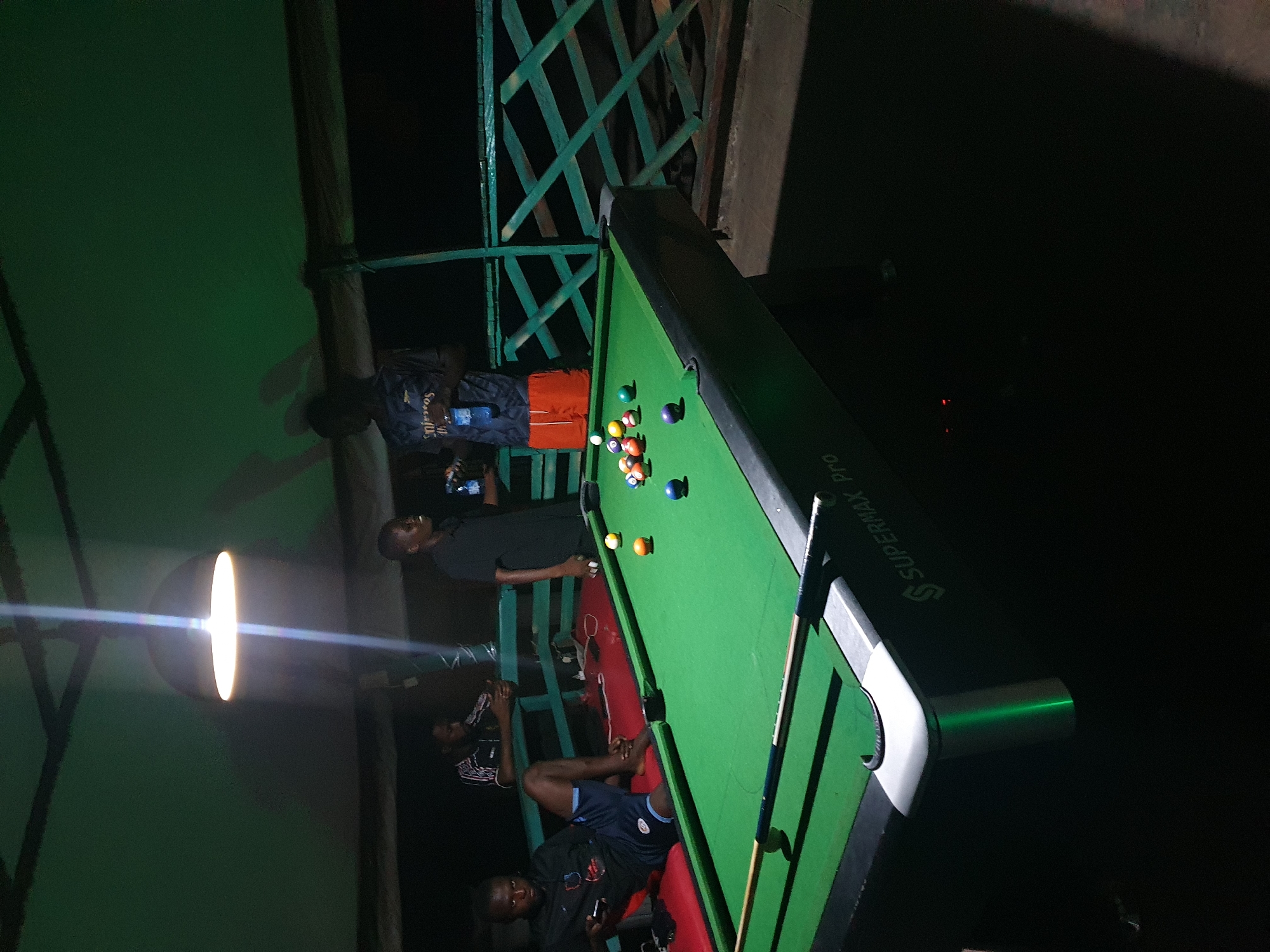 The image depicts an outdoor setting at night, likely a bar or recreational area, with a billiard table as the central focus. Three men are visible. One man in the foreground, seated on a red bench to the left of the billiard table, is looking towards the viewer while holding a mobile phone. He is wearing a dark t-shirt with a crest and dark shorts with a light-colored trim. Another man stands behind the billiard table on the right, facing a third man who is also standing and holding a mobile phone. This second man is wearing a dark t-shirt and orange shorts. The third man, positioned further back and to the left, is seated and wearing a patterned t-shirt. The billiard table is green with visible balls arranged in a typical setup. A pool cue rests on the table. The background features a green tarp or fabric overhead, supported by dark beams, and a green latticed railing. The lighting appears to be artificial, with a prominent overhead lamp casting a bright glow. The overall ambiance suggests a casual, social gathering. The scene is situated in Lokoja, Nigeria, an urban environment at night.