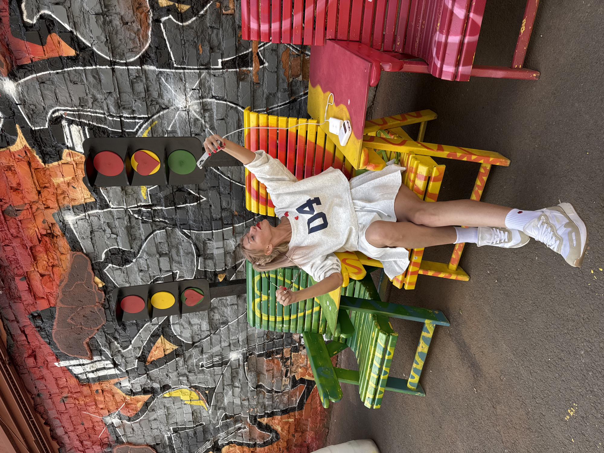 A blonde woman in a white sweater and skirt is sitting on a colorful chair and taking a selfie with her phone, which is connected to a power bank. She is in front of a graffiti-covered brick wall with a traffic light installation that has a red light, a yellow light, and a heart-shaped green light. The chairs are painted in bright colors, including green, yellow, and red, and have a whimsical design. The woman is wearing white sneakers and white socks. The location is likely an outdoor art installation or a public space in Saint Petersburg, Russia, given the context of the provided crops showing graffiti and colorful street art.