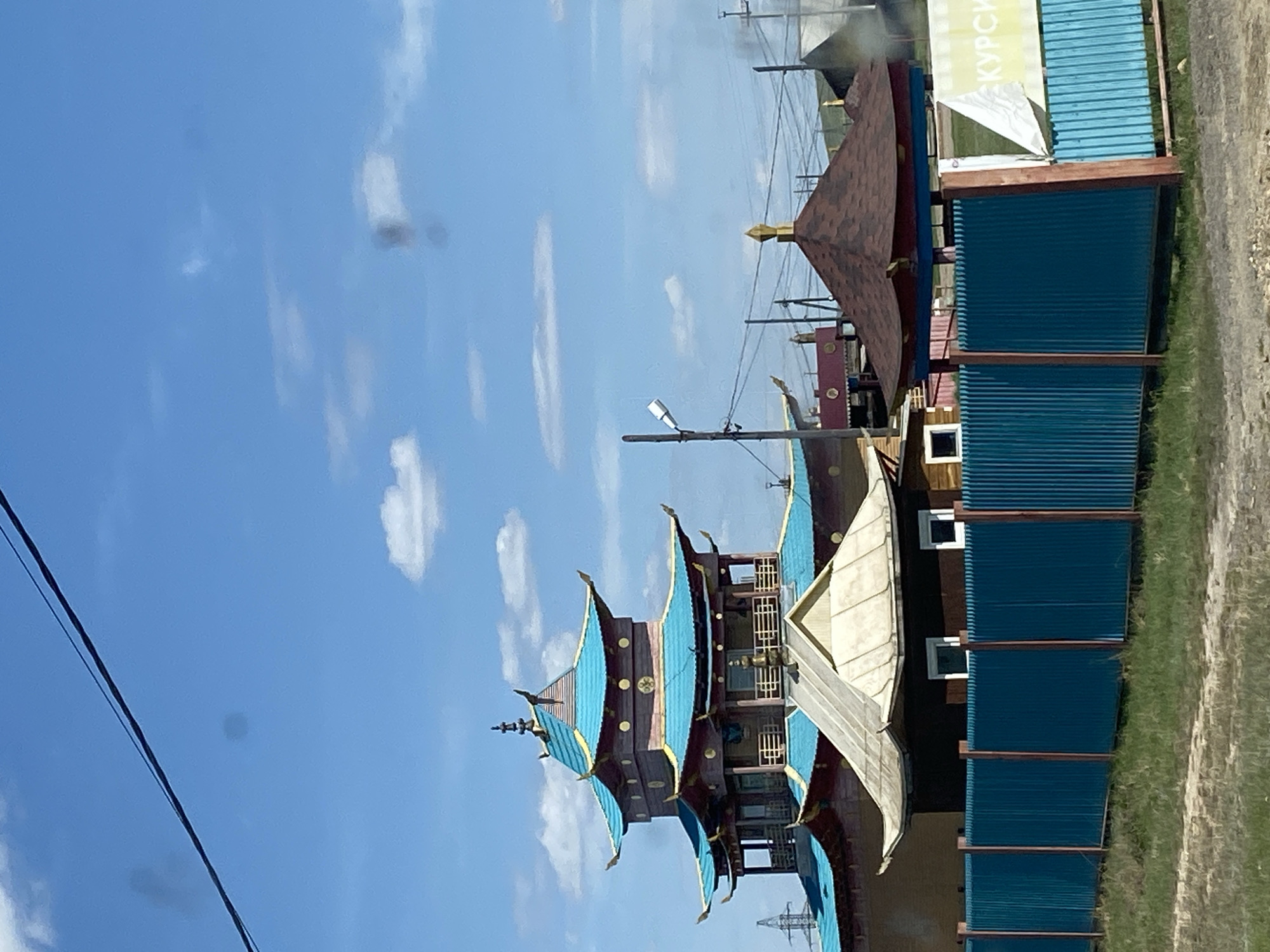 Here is a description of the image:
The image shows a vibrant and detailed architectural structure, seemingly a multi-tiered pagoda or tower, characterized by its blue and brown color scheme and ornate design elements. The architecture appears to be influenced by Asian styles, possibly Chinese or Japanese. This structure is situated next to what seems to be a modern, utilitarian building, which is constructed from blue corrugated metal sheets. The ground surrounding the buildings features green vegetation.
In the background, a clear blue sky dotted with scattered white clouds is visible. There are power lines running across the sky, suggesting the structures are situated in a developed area.
Based on the information available, this image likely depicts a site in Ust-Ordynsky, Russia. The ornate pagoda structure stands in contrast to the more functional, modern structure beside it.