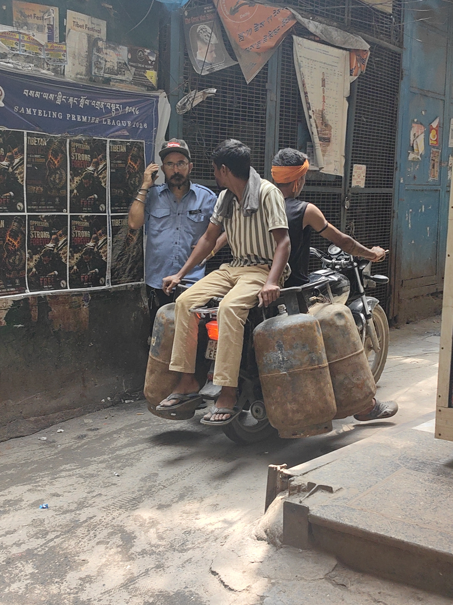 In Delhi, India, on a street with rough asphalt and debris, two men are riding a motorcycle loaded with two large, rusty gas cylinders secured to the back. The man in front, wearing a blue shirt and a cap, is looking towards his right while holding his left hand to his ear, possibly on a phone. The man behind him, with an orange headband and a striped shirt, is looking ahead, driving the motorcycle. The cylinders are positioned vertically on either side of the rear wheel, with one cylinder slightly in front of the other. In the background, posters advertising "Tibet Festival" and "SAMYELING PREMIER LEAGUE 2026" are visible on a wall, along with a blue metal gate and wire fencing. The scene appears to be captured during daylight.