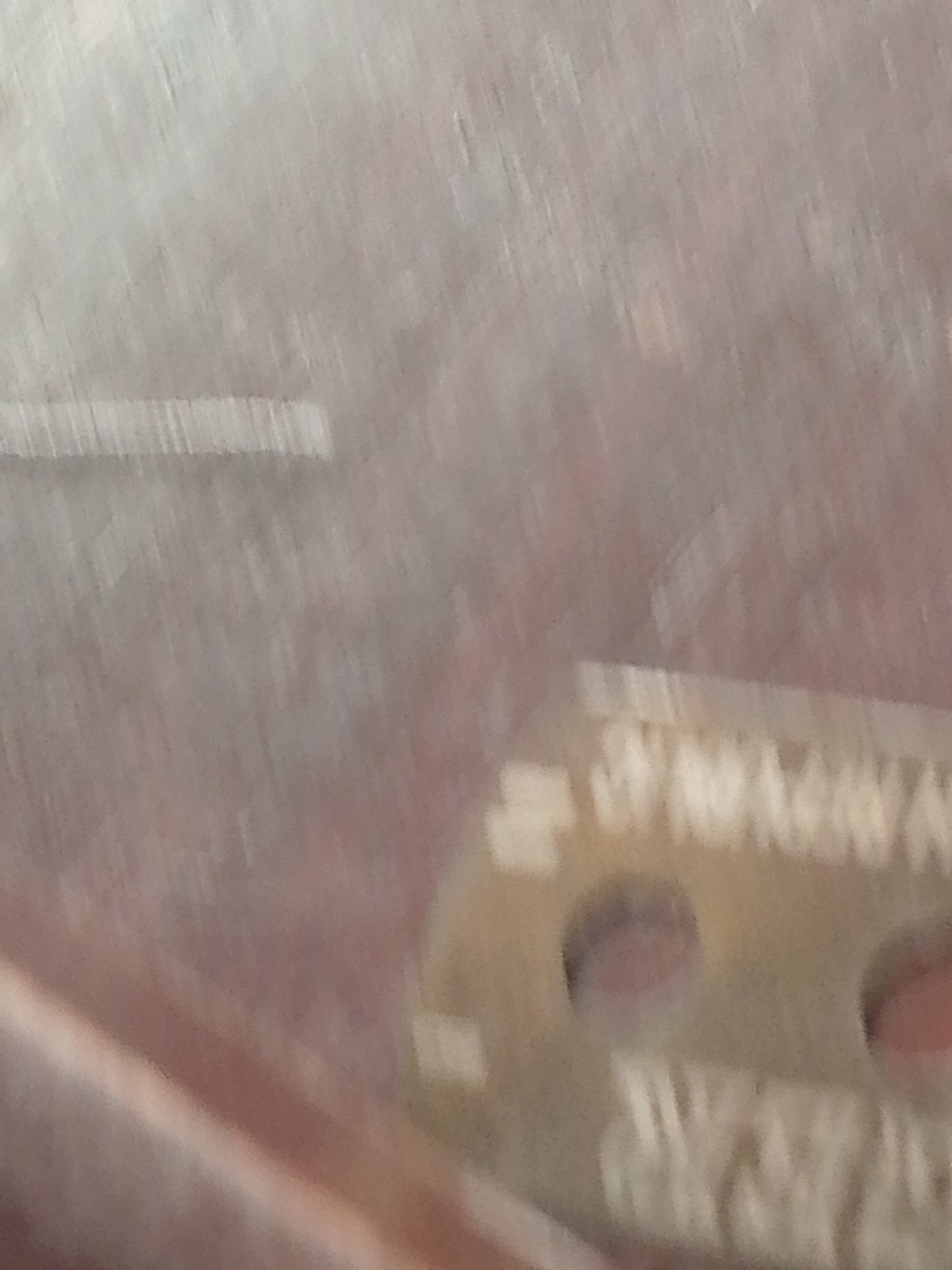 This is a close-up, out-of-focus image of a metallic object, possibly a tool or a part of a machine, with visible text that is also blurred. The background appears to be a rough, textured surface with a reddish-brown hue, possibly metal or concrete. There is a faint horizontal line across the upper left of the image. The metallic object has a series of raised ridges and circular indentations. Text is present on this object, appearing as repeated patterns of what could be letters, but is illegible due to the blur. The image does not contain any discernible elements that would allow for localization within Jauro Yinu, Nigeria, or any specific urban or environmental context.