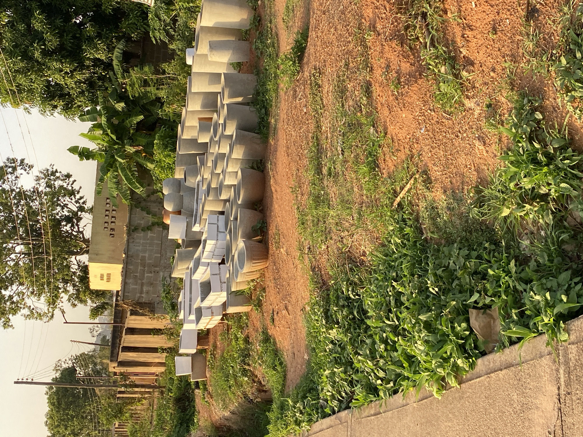 The scene depicts a roadside area in Bukuru, Nigeria. A collection of cylindrical concrete pots, possibly for plants or landscaping, are stacked along the roadside. Behind the pots, there is a building with several windows and a section with what appears to be ventilation blocks. The ground is covered with vegetation, including various grasses and small plants. The overall environment suggests an urban or suburban setting within the city.