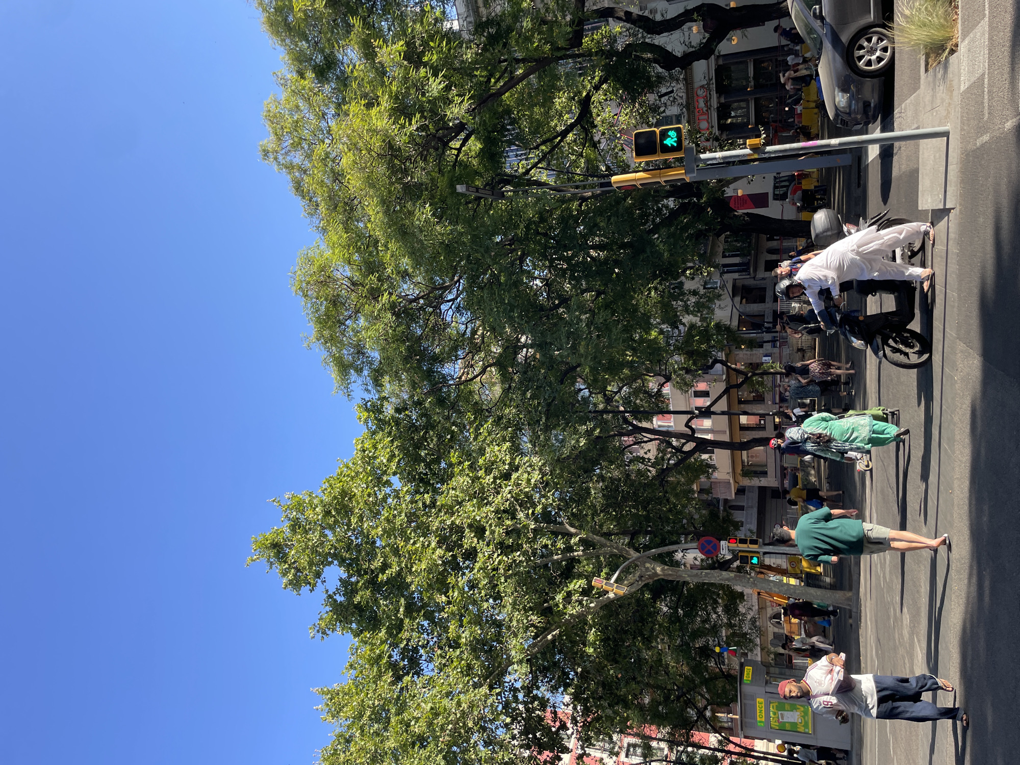 This image depicts a street scene in Barcelona, Spain, on a sunny day with a clear blue sky. In the foreground, several people are walking across a street. A man in a white shirt with red accents and a red cap is walking from left to right, carrying a pink folded item. To his right, a man in a green t-shirt and grey shorts is walking away from the camera. A woman in a green traditional garment is walking from right to left, pulling a green cart. On the right side of the image, a man dressed in white traditional clothing is sitting on a black scooter, facing left. A traffic light for pedestrians is visible, showing a green walking figure. A gray car is parked on the right side of the street, with a building behind it. Large, leafy trees line the street, casting shadows. Buildings with visible windows and balconies are present in the background. A yellow kiosk with "ONCE" signage is on the left side of the image. The scene suggests a bustling urban environment.