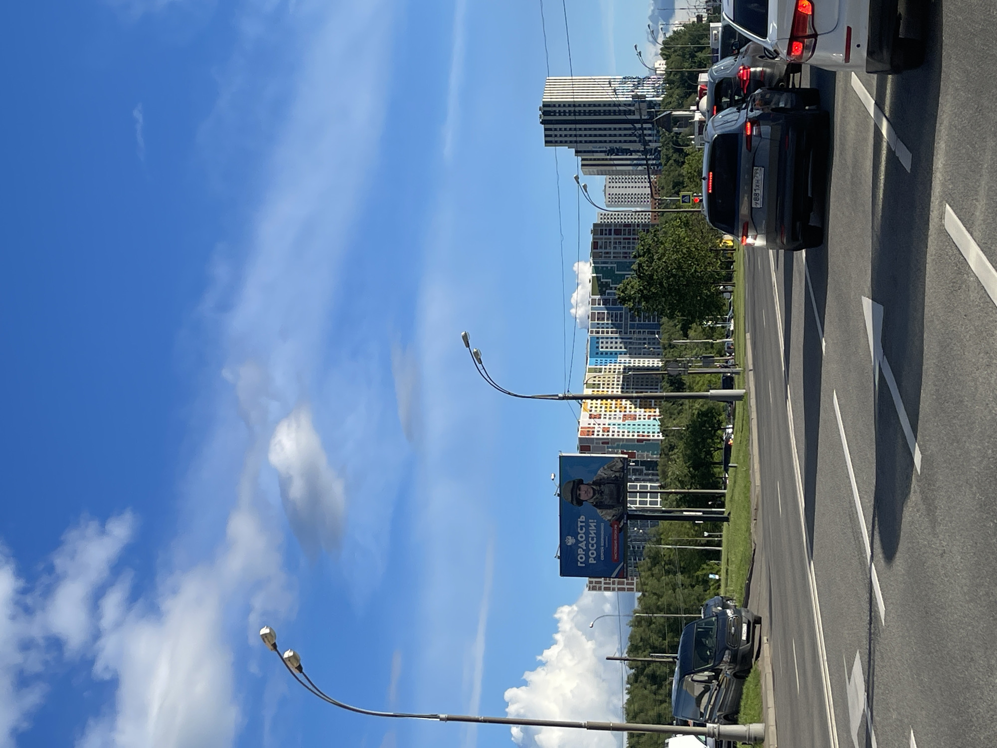 The image shows a street in Moscow, Russia. The view is along a multilane road, with buildings visible in the background. Traffic is heavy, and cars are visible in the foreground and middle distance. A large billboard is seen on the left side of the road. Streetlights line the road. The sky is blue with scattered clouds.
