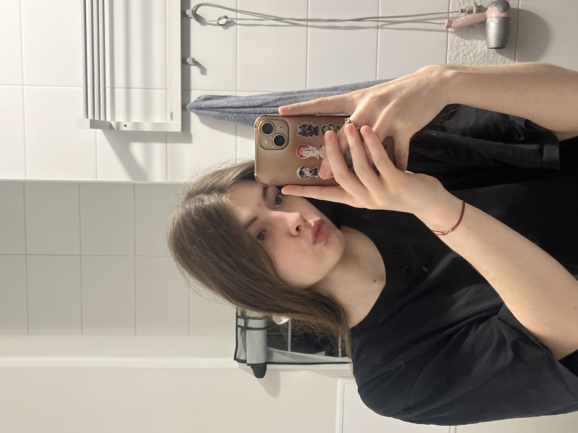 A young woman is taking a selfie in a bathroom. The walls are tiled in white. In the background, a heated towel rail and a hairdryer are visible. The woman is holding a phone with a brown case decorated with stickers. She has light brown hair and is wearing a black t-shirt. There is a red bracelet on her left wrist. There is no information to suggest a location in the city of Saint Petersburg, Russia.