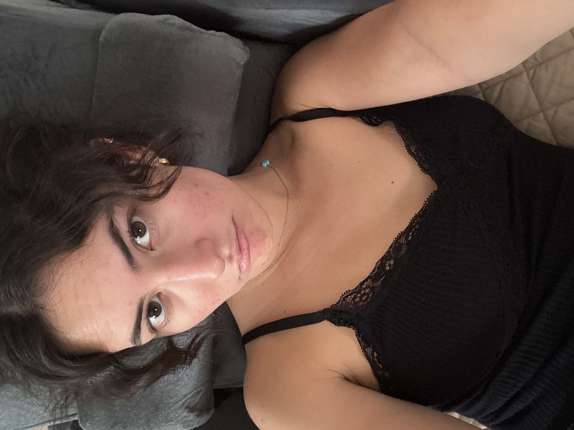 A young woman is lying on a gray couch. She is wearing a black, lacy top. She has brown eyes and dark curly hair. A light blue pendant is visible on her necklace. The background includes a quilted beige blanket or sheet. The location is unknown and there is no indication in the image it is in the city of Puebla, Mexico.