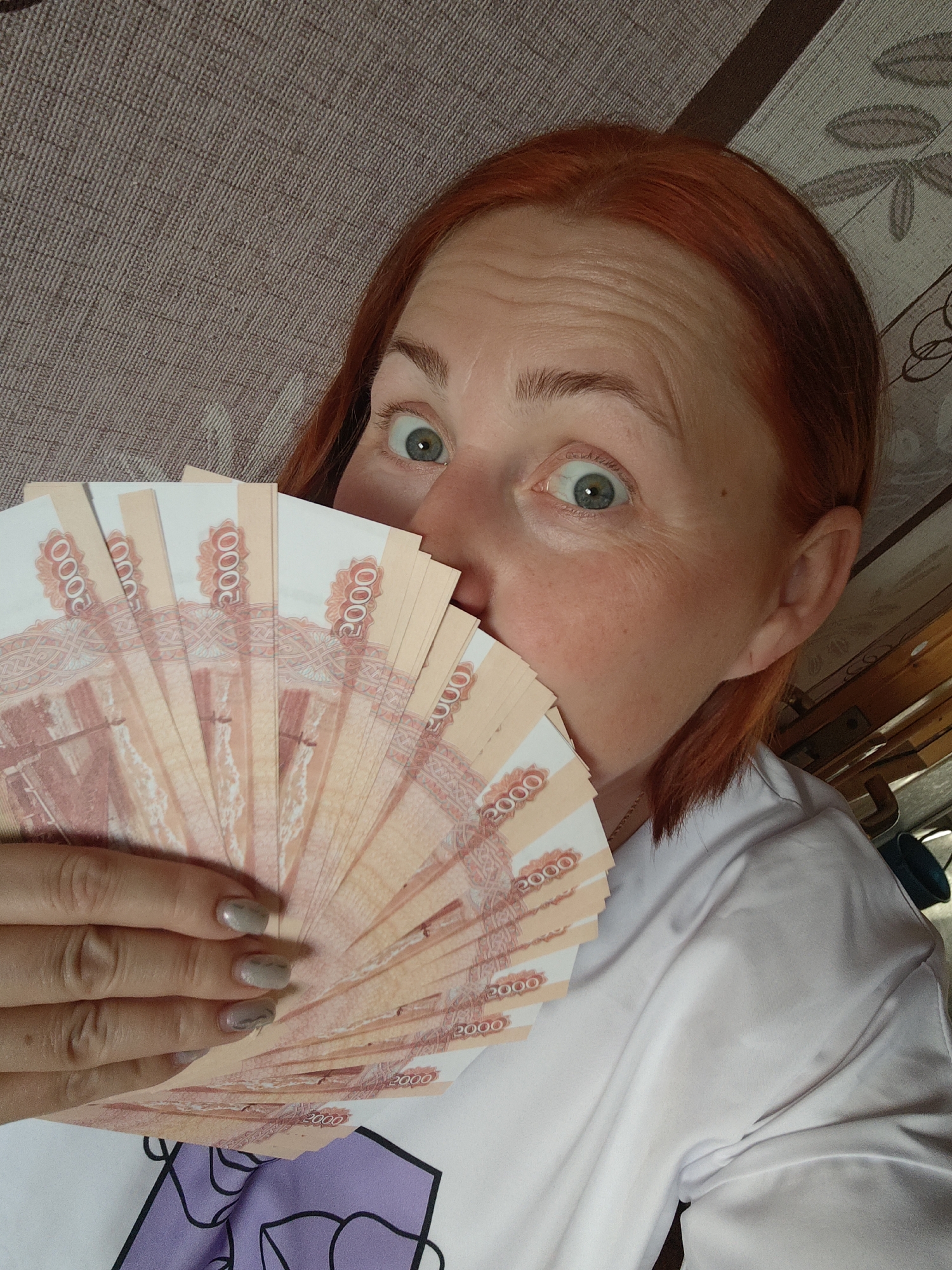 A fair-skinned woman with red hair is holding a large fan of 2000 Russian ruble notes in front of her face, obscuring her mouth and nose. Her eyes are wide with an expression of surprise or excitement. She is wearing a white t-shirt. The background includes a textured surface, possibly a rug or blanket. The location is Марковский, Russia.