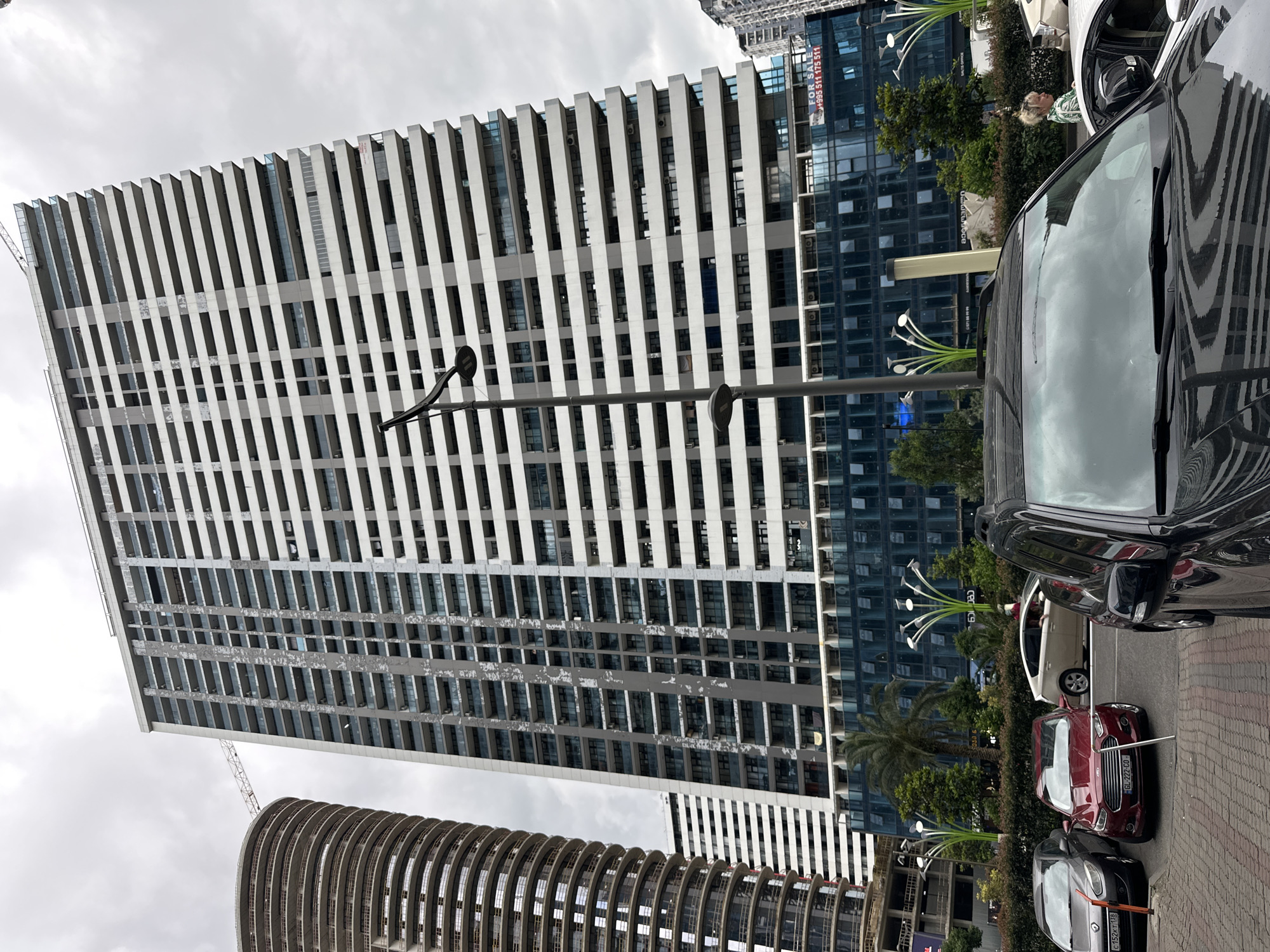 The image shows a wide, low-angle exterior view of a very tall, modern apartment building in Batumi, Georgia. The building features a white facade with alternating grey horizontal bands and numerous balconies. To the left of this building is a partially constructed, cylindrical tower under construction, with exposed rebar and concrete floors. In the foreground, several cars are parked, including a dark SUV in the immediate foreground, a red sedan, a dark grey sedan, and a light-colored hybrid car. A woman with blonde hair pulled into a bun, wearing a green and white patterned shirt, is walking past the cars. The street in front of the buildings has patterned brick paving. Decorative streetlights with a futuristic, bulbous design are visible in the middle ground, flanking a palm tree. A large banner with the words "FOR SALE" and a phone number is affixed to the lower section of the apartment building. The sky is overcast and cloudy. The scene appears to be a street in a contemporary urban area of Batumi, likely a district with ongoing or recent development.