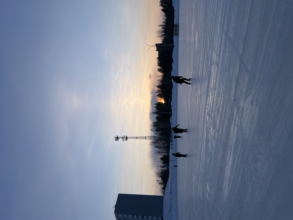 The image displays an outdoor ice skating rink in Ust-Ilimsk, Russia, captured during either a sunrise or sunset. Five individuals, rendered in silhouette, are actively ice skating on the expansive ice surface. The foreground is dominated by the textured, blue-tinged ice. In the background, a tall, multi-story dark building occupies the far left. A prominent, lattice-style metal communication tower stands to its right. A dense line of dark, snow-dusted coniferous trees forms the horizon line, behind which a distinct orange and yellow glow is visible, indicative of the sun's low position or a distant light source. Snow covers the ground beyond the rink, where a street light pole and what appear to be benches are also visible on the right.