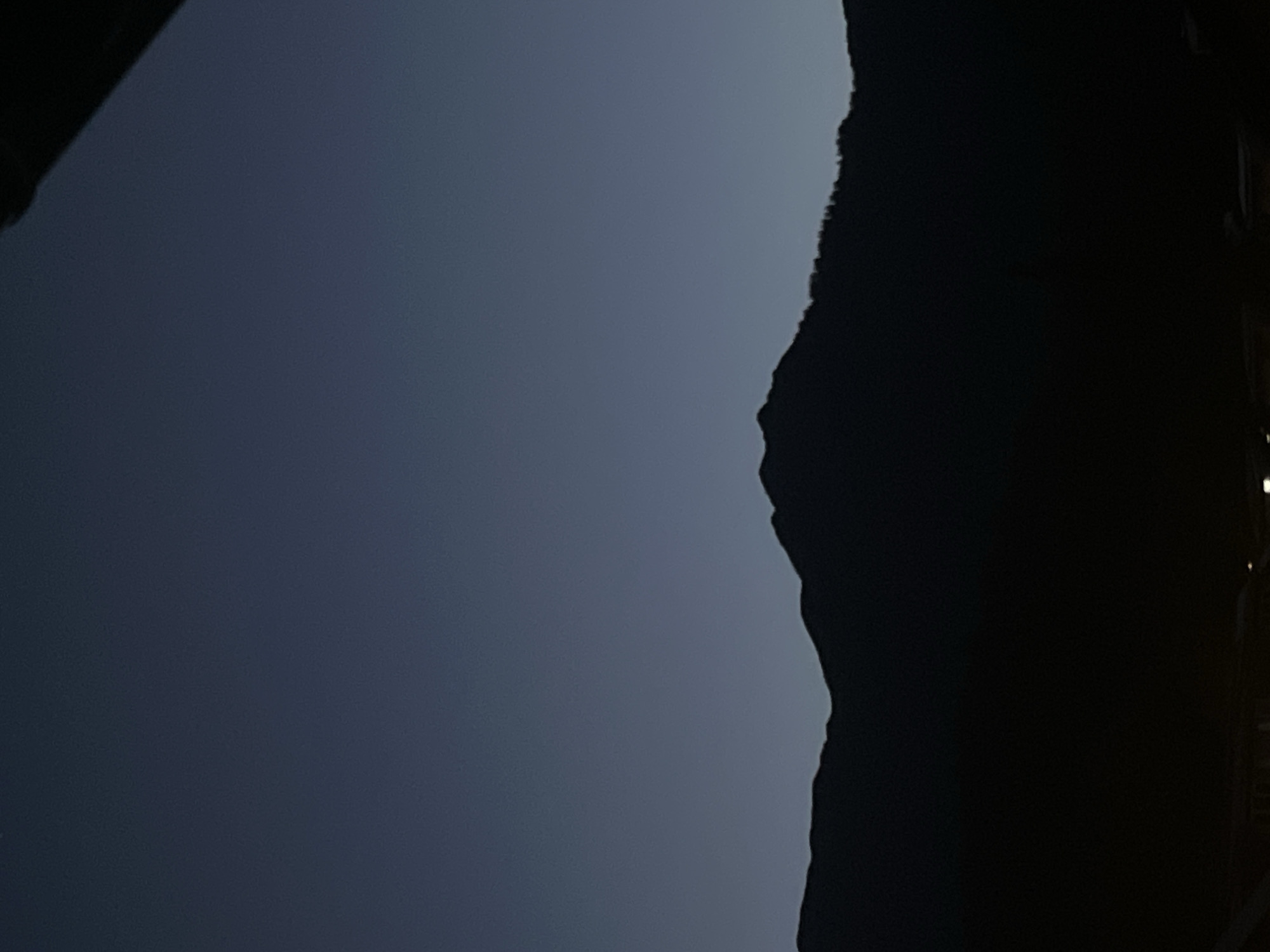 This image depicts a silhouetted mountain range against a dark, twilight sky, likely taken in Garmisch-Partenkirchen, Germany. The mountains are dark and indistinct, with a slightly jagged treeline visible along the upper slopes. The sky is a deep, gradient blue, transitioning to a lighter hue near the horizon, suggesting the last light of day or the first light of dawn. In the foreground, at the bottom of the frame, there are faint lights that could indicate a town or settlement nestled at the base of the mountains. The overall impression is one of a serene, natural landscape at dusk or dawn.