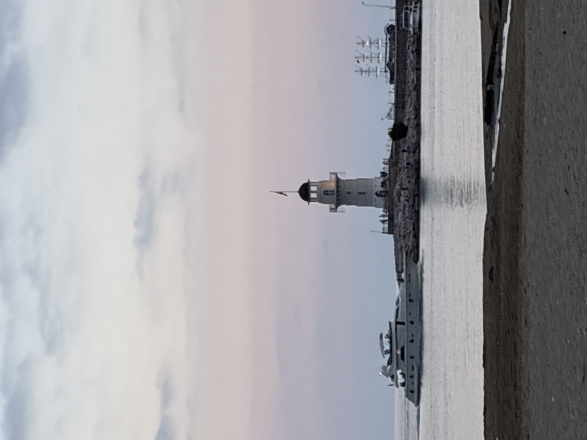 This image presents a tranquil coastal scene, likely a harbor entrance or marina, set against a soft, diffused light indicative of either early morning or late evening twilight. The primary subject is a white lighthouse, prominently positioned towards the left-center of the frame, featuring a distinct black domed cap and a small flag fluttering from its peak.
To the right of the lighthouse, a sleek, white luxury yacht or large boat floats calmly in the water, displaying multiple decks and windows that catch the gentle light. The water surface is relatively placid, exhibiting subtle reflections. A rocky breakwater or pier extends from the right, providing a boundary between the land and the sea, with some indistinct structures, possibly part of a marina or port, visible further in the background.
The sky above is partly cloudy, painted with delicate pastel shades of light blue, soft white, and faint pinkish-purple, creating a peaceful ambiance. The immediate foreground on the right reveals a textured concrete or stone surface, suggesting the photograph was taken from another structure overlooking the water. No specific activities or individuals are discernible, lending a quiet and still quality to the scene. No legible text is visible anywhere in the image. The setting is consistent with a coastal port in Alanya, Turkey.