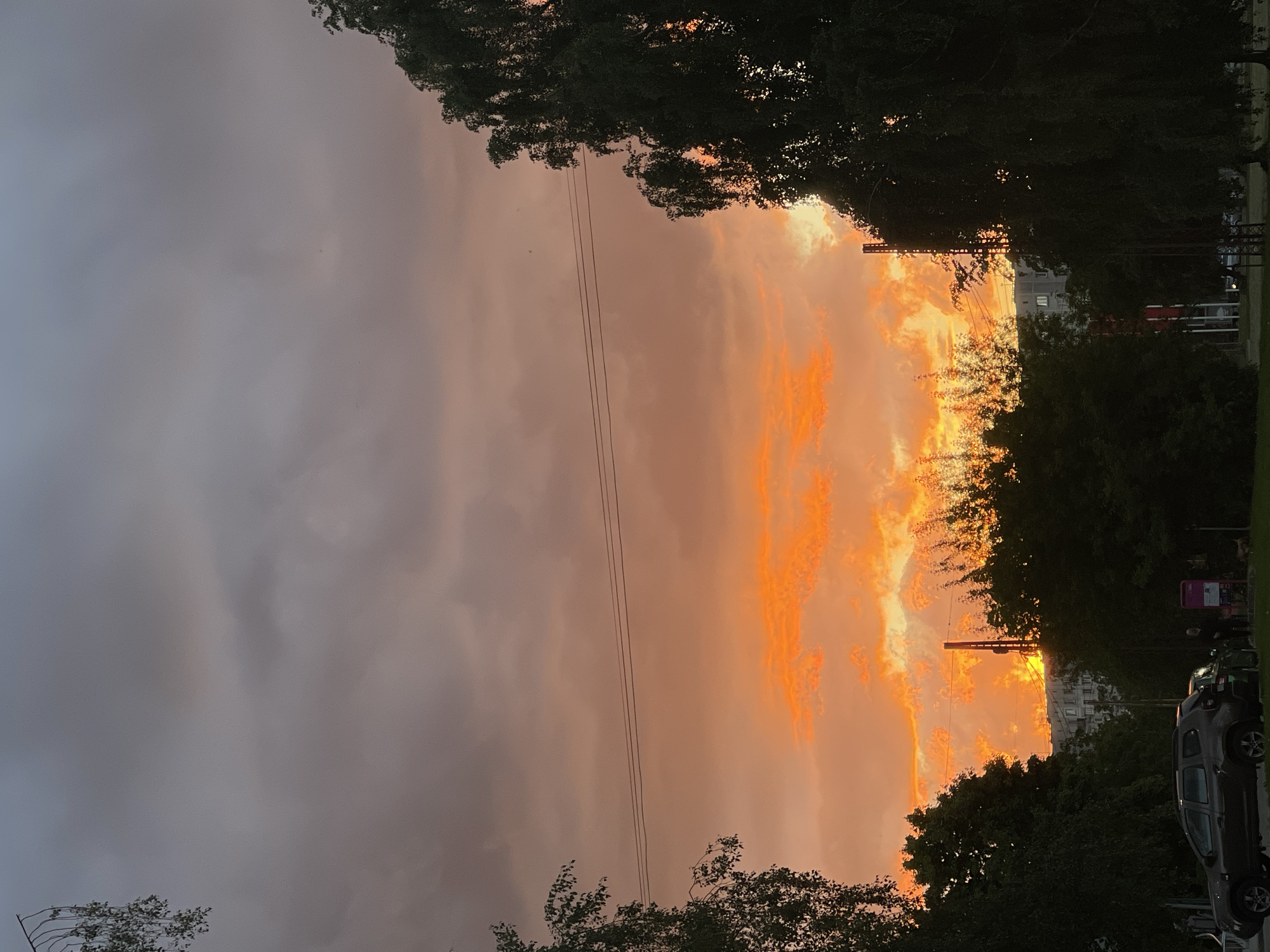 The image captures a vivid sunset in Riga, Latvia. The sky transitions from a dark, overcast gray on the left to a fiery orange and yellow on the right, indicative of the setting sun. The sunset is partially obscured by trees lining what appears to be a street or avenue. Electrical lines run horizontally across the middle of the image. In the lower right, several cars are parked along the curb. A building is visible through the trees in the middle-right.