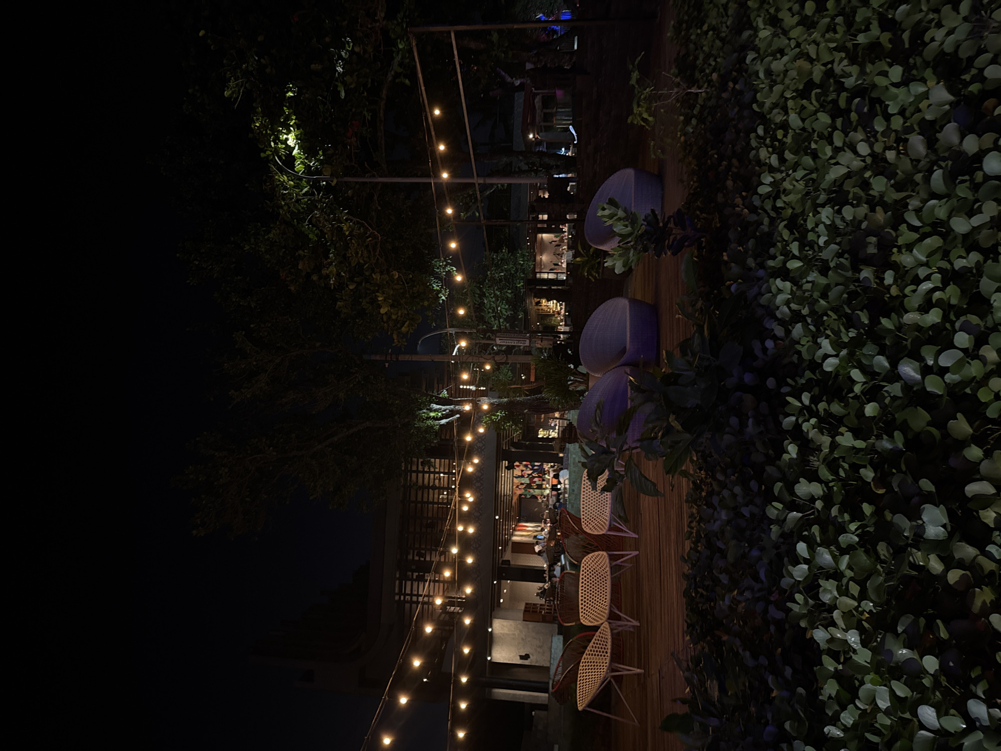 The image captures an outdoor seating area at night, likely a restaurant or cafe, in Kuta, Indonesia. String lights illuminate the scene, casting a warm glow over a wooden deck furnished with several white woven chairs and two large, purple, rounded lounge chairs. In the background, a multi-story building is visible with open-air sections and more illuminated string lights, suggesting an indoor dining area with patrons. Trees and lush green ground cover surround the seating area, with a street lamp visible to the right, its light partially illuminating the foliage. The overall atmosphere is serene and inviting, indicative of a popular nighttime venue.