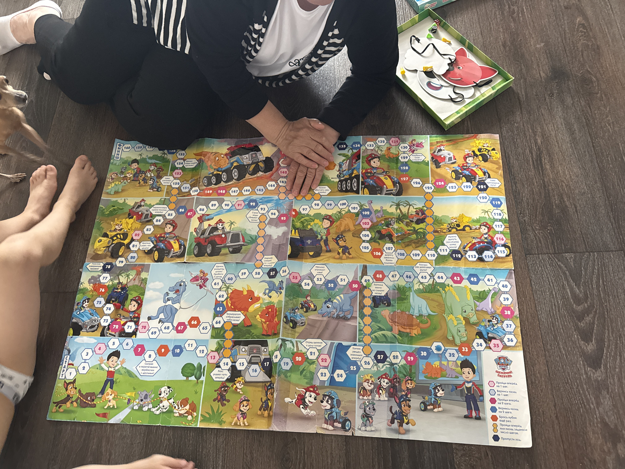 The image shows a top-down view of a Paw Patrol board game laid out on a wooden floor. The board game features colorful illustrations of characters and vehicles from the Paw Patrol franchise, with numbered spaces indicating game progress. Several people are partially visible in the frame, including a child's legs and feet on the left, and an adult sitting on the right with their legs crossed and wearing black pants and a black and white striped shirt. A small tan dog is also visible in the upper portion of the image, appearing to be standing or walking. To the right of the board game, a green box with a cutout shape containing game pieces is visible. The overall scene suggests a recreational activity taking place indoors. The image does not contain any information that would allow for the identification of the specific location within the city of Chaikovsky, Russia.