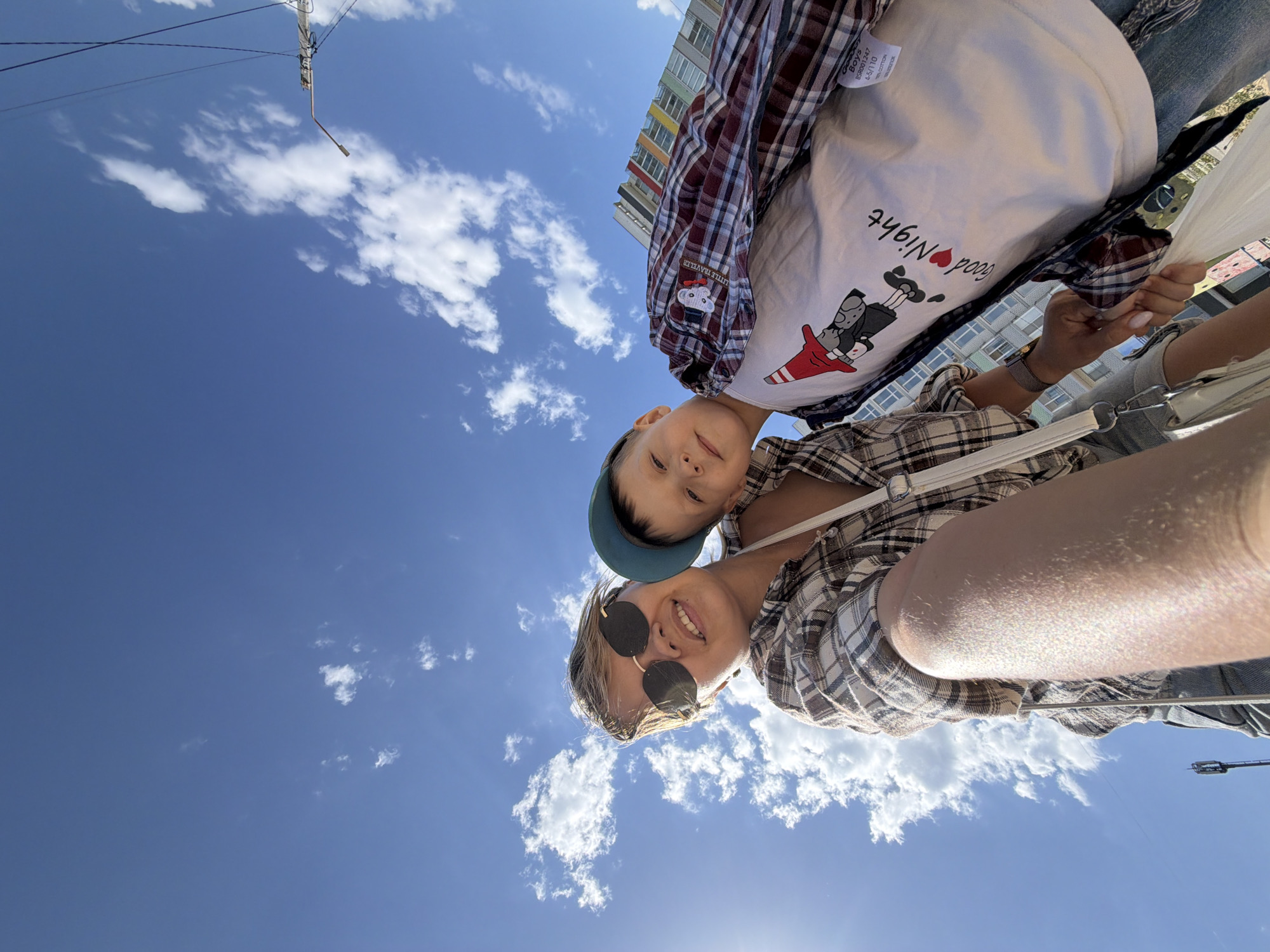 A selfie looking upward features a woman with sunglasses and a young boy with a cap. The woman wears a plaid shirt, and the boy wears a shirt with a "Good Night" graphic and a plaid jacket. The background is a bright blue sky with scattered white clouds. An electrical pole with wires is partially visible in the top left corner. The photo appears to be taken in Chaikovsky, Russia.