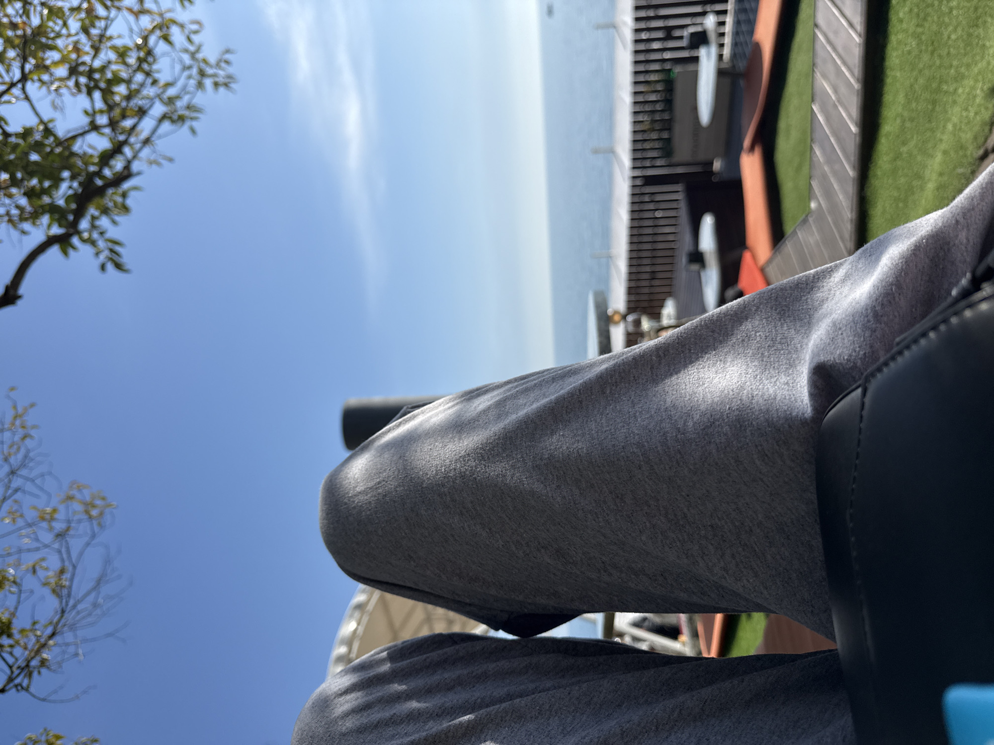 The image captures an outdoor scene from a first-person perspective, looking out over a wide body of water on a clear, sunny day. The primary subject is the lower half of a person's body, specifically their legs, dressed in grey sweatpants. The individual appears to be seated in a dark, possibly leather, chair or lounge, visible on the right side of the frame.
The setting is an elevated outdoor area, likely a deck, balcony, or rooftop, providing a panoramic view. Beyond the foreground elements, a vast expanse of calm, light blue water stretches towards the horizon, suggesting an ocean or a large lake. The distant horizon where the water meets the sky is slightly hazy. The sky above is a bright, clear blue, adorned with a few delicate, wispy white clouds.
In the upper left corner, a tree branch with green foliage is visible, appearing slightly out of focus. The immediate surroundings of the seating area include what looks like dark wooden decking and patches of artificial green turf or grass, hinting at a structured outdoor leisure space. Bright sunlight illuminates the scene, casting soft shadows on the person's clothing and highlighting the texture of the fabric. The overall atmosphere conveyed is one of relaxation and peace, enjoying a scenic view under pleasant weather conditions. No discernible text is visible within the image.